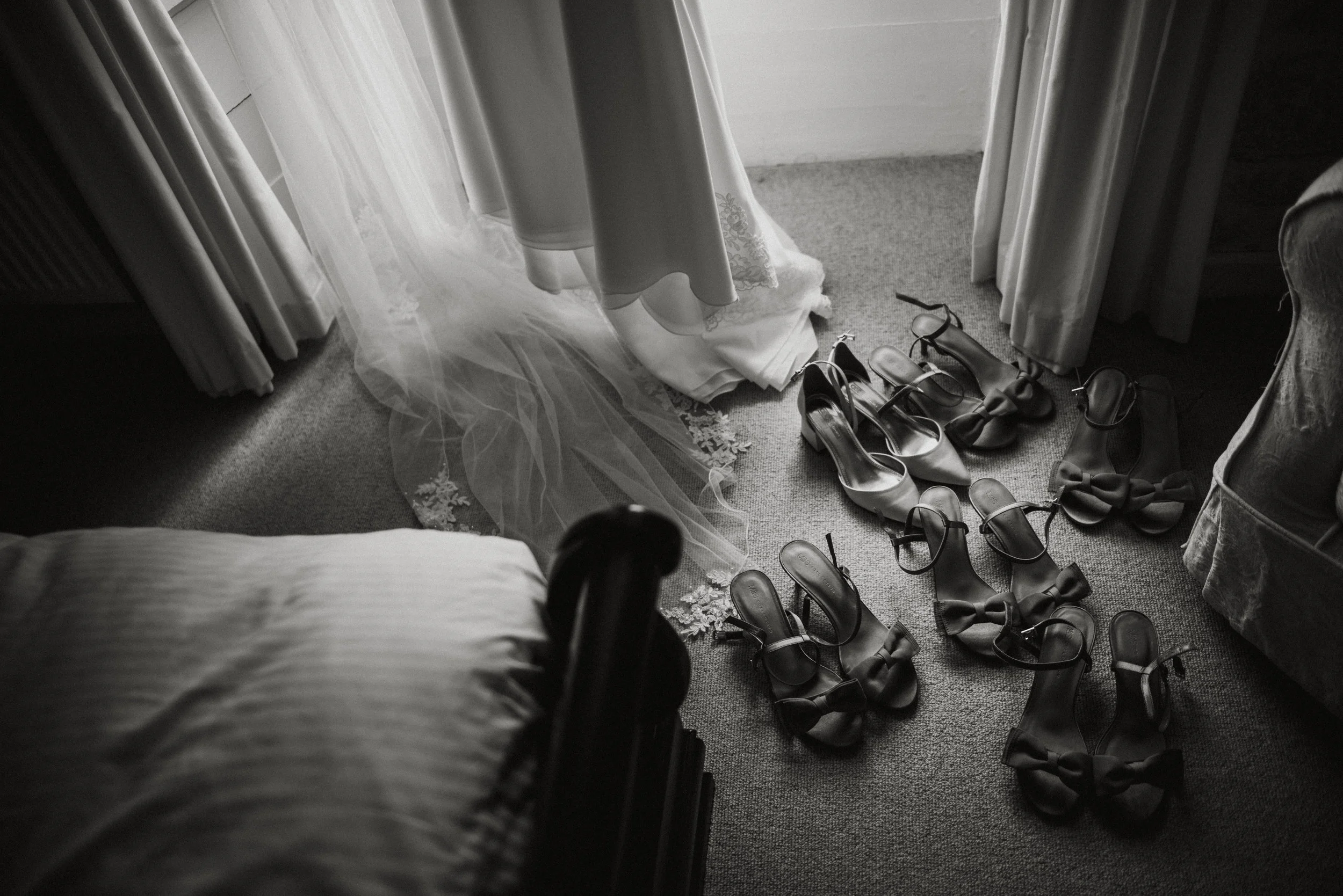 Multiple pairs of high-heeled shoes and a wedding dress on the floor near a bed and curtains in a monochrome room.