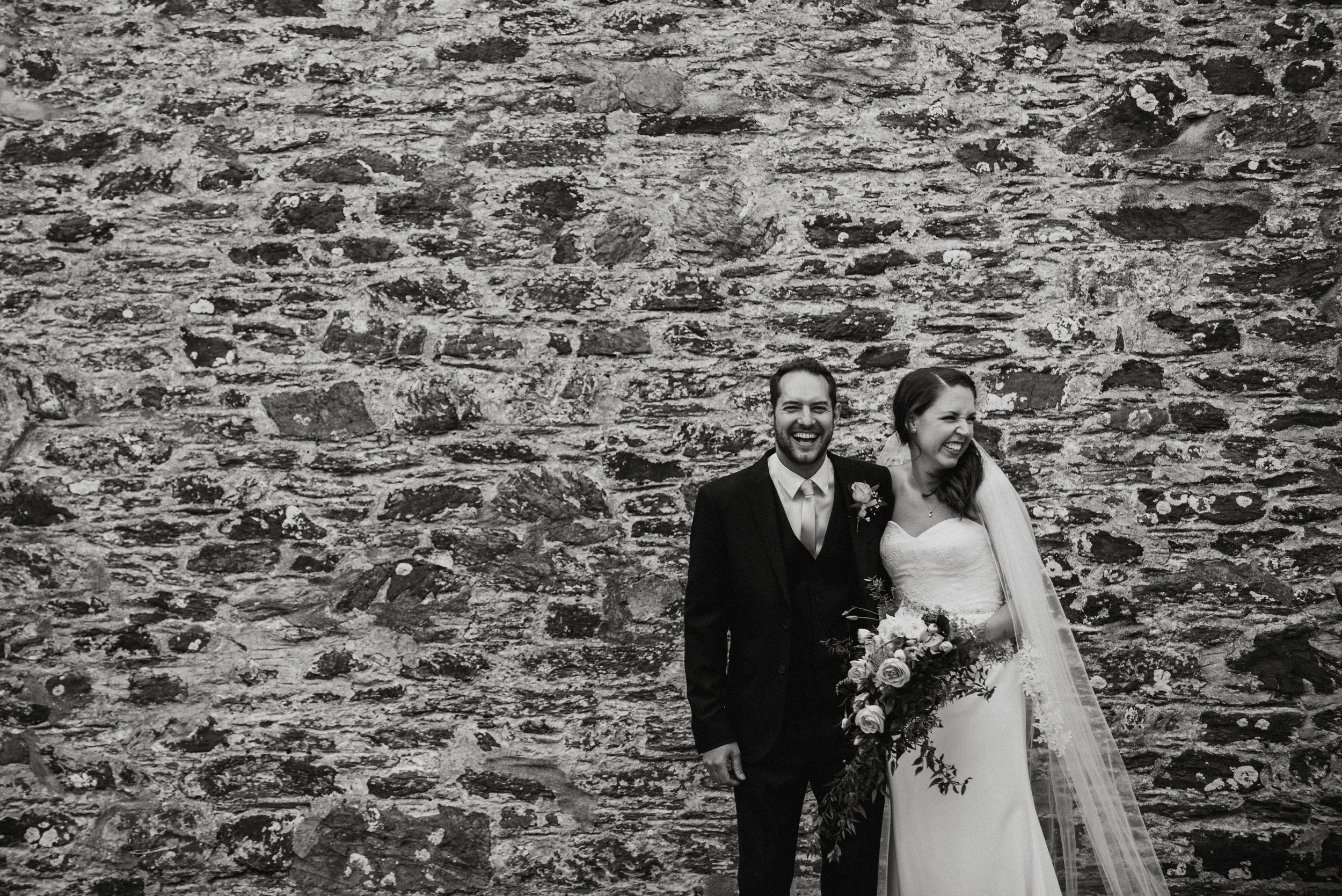 Black and white photo of a happy bride and groom standing in front of a stone wall. The bride is holding a large bouquet of flowers, and both are smiling and laughing.