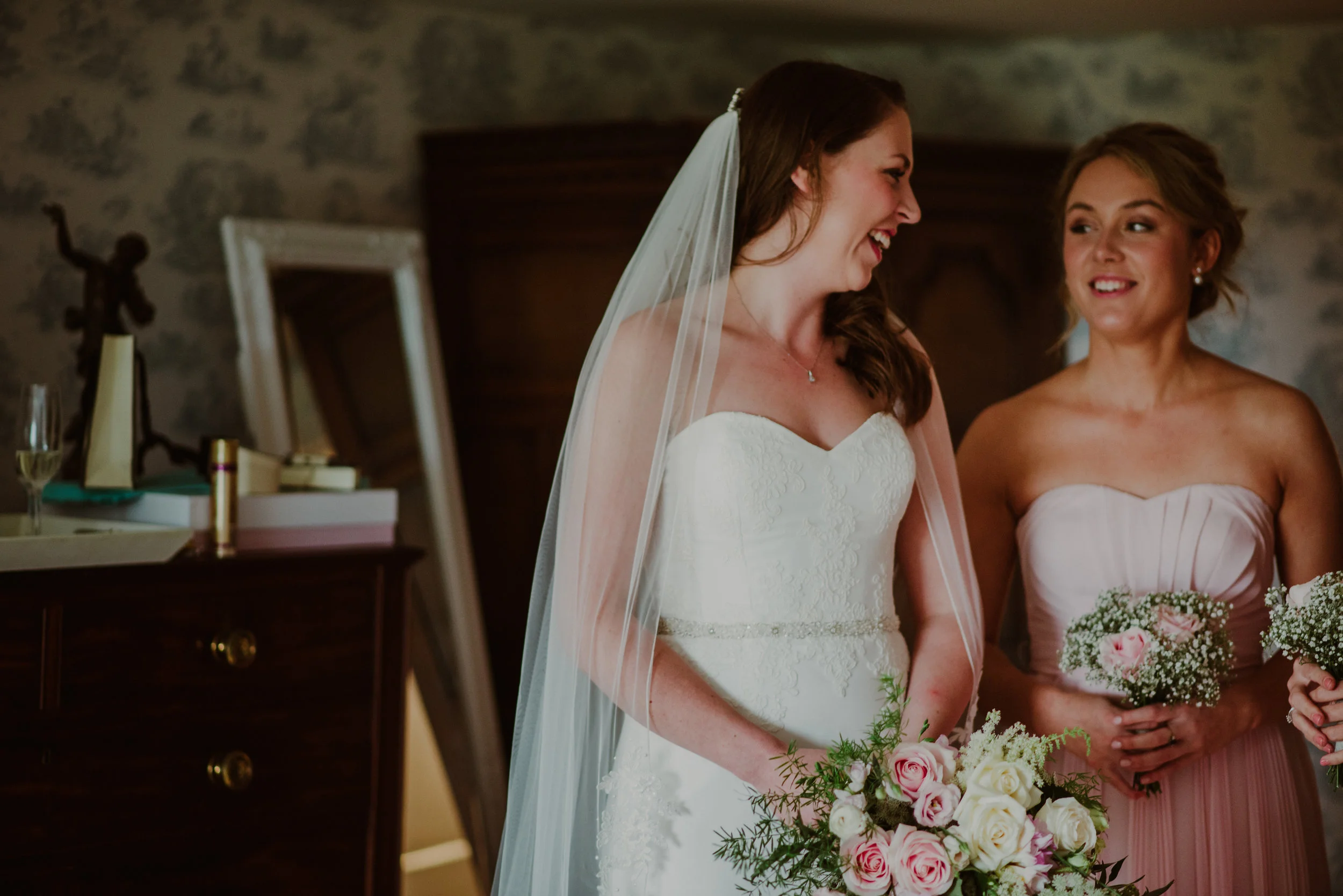 A bride in a white lace wedding dress and veil, holding a bouquet of pink and white roses, smiling and talking with a bridesmaid in a strapless pink gown holding a smaller bouquet, in a warmly lit indoor setting.