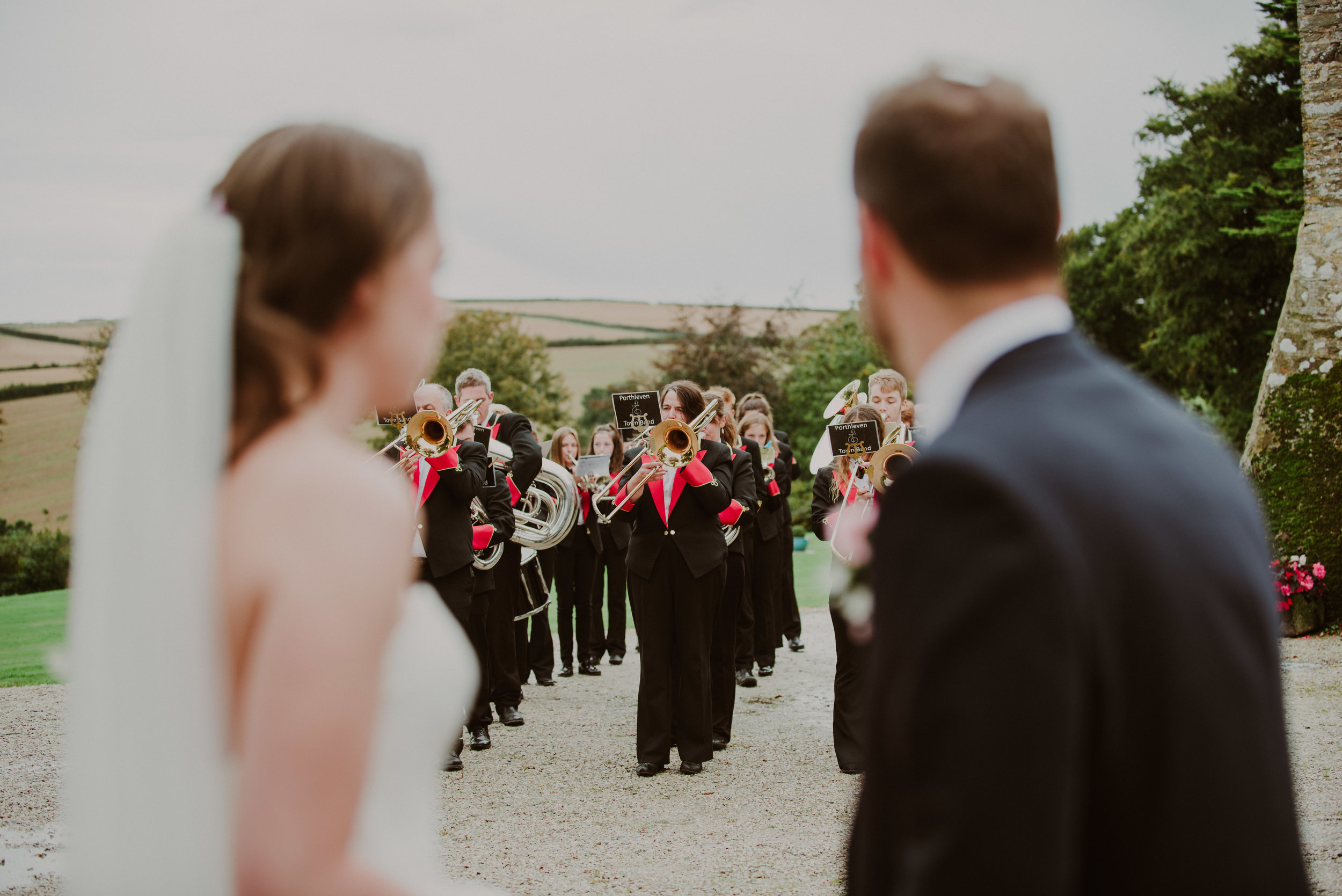 Wedding couple standing for a photo before a brass band performance outdoors, with a scenic background of fields and trees.
