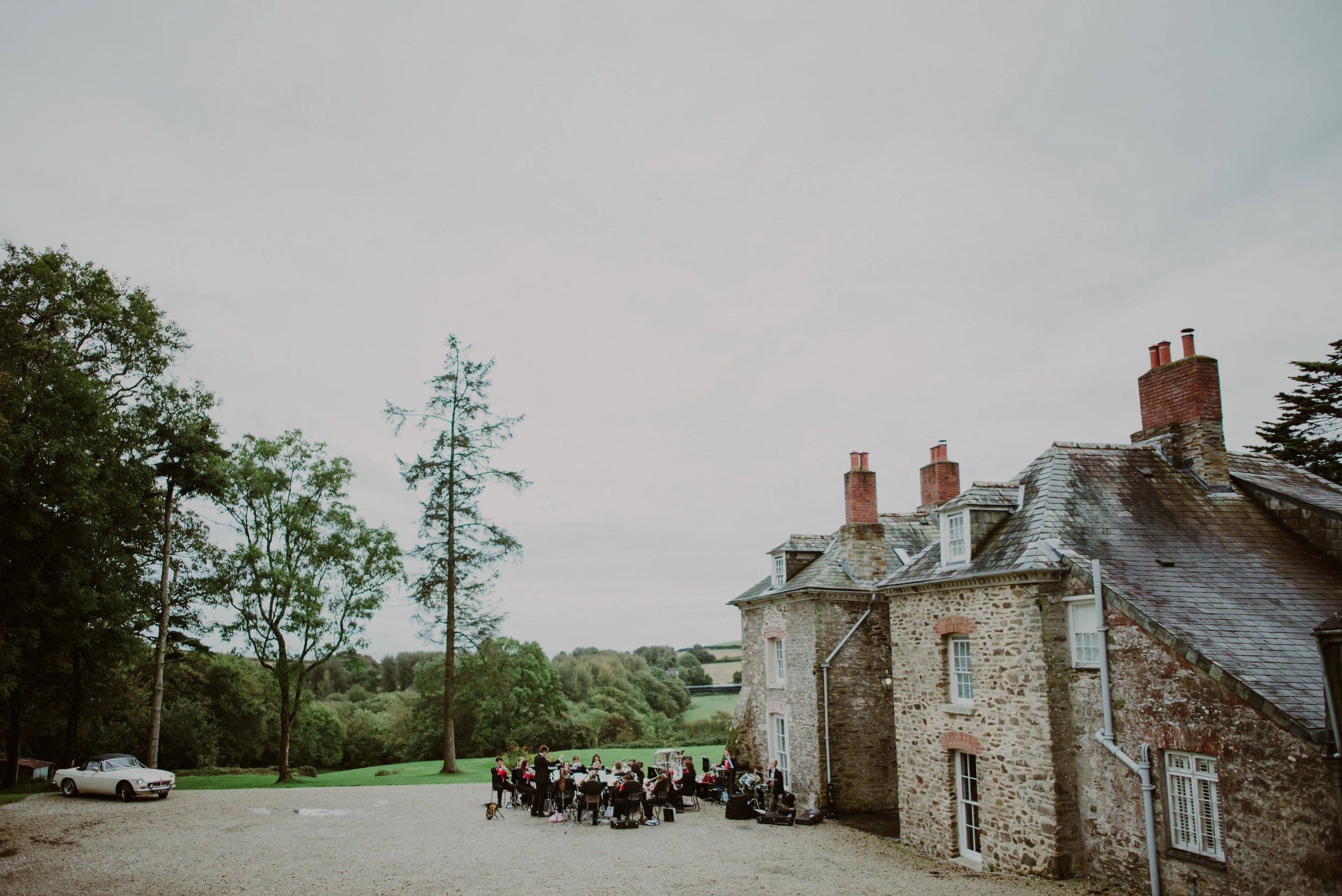 An outdoor scene at a large stone house with a group of musicians performing in the gravel yard, surrounded by trees and rolling hills.