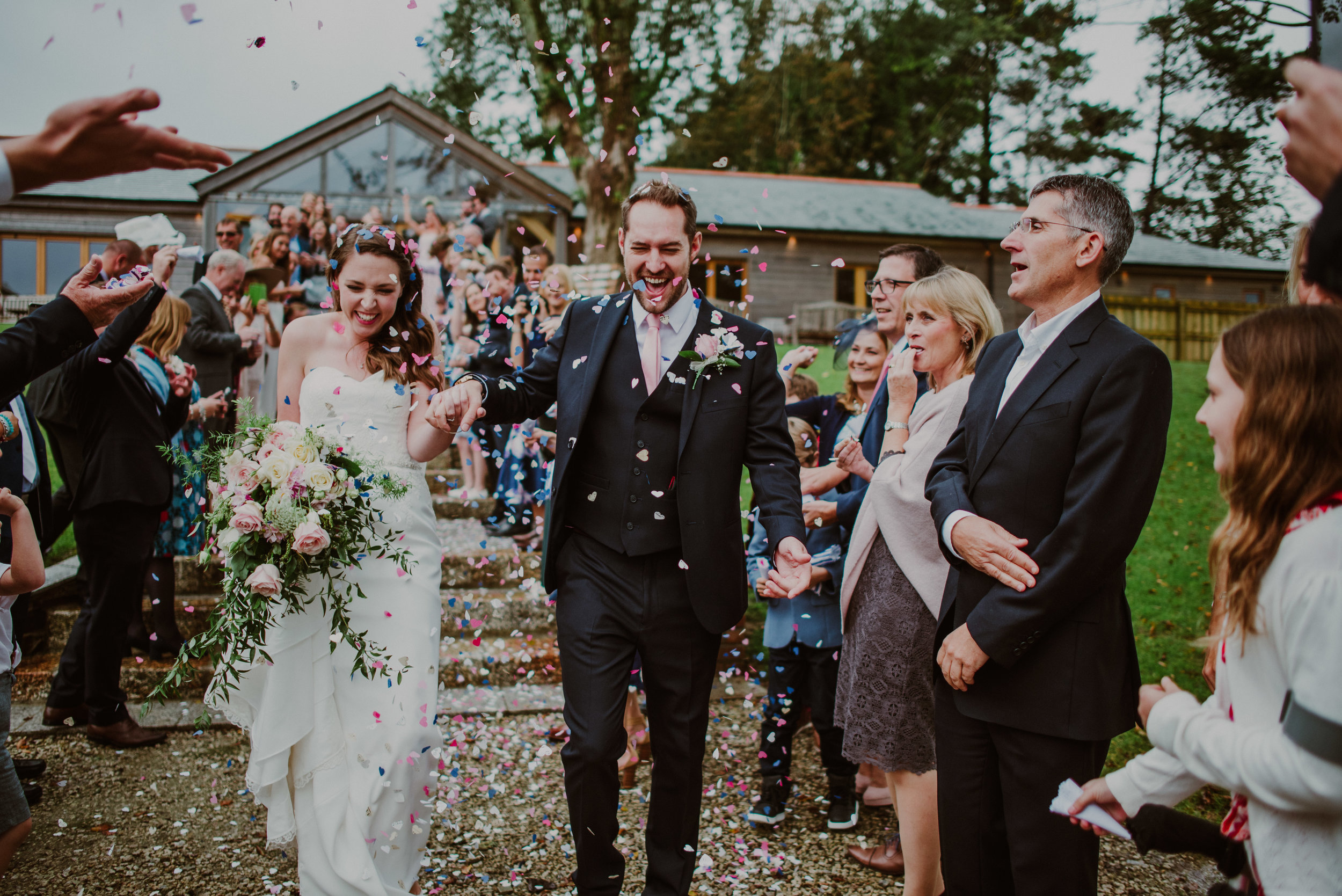 A newlywed couple walking hand-in-hand through a shower of colorful confetti, surrounded by smiling friends and family outdoors.