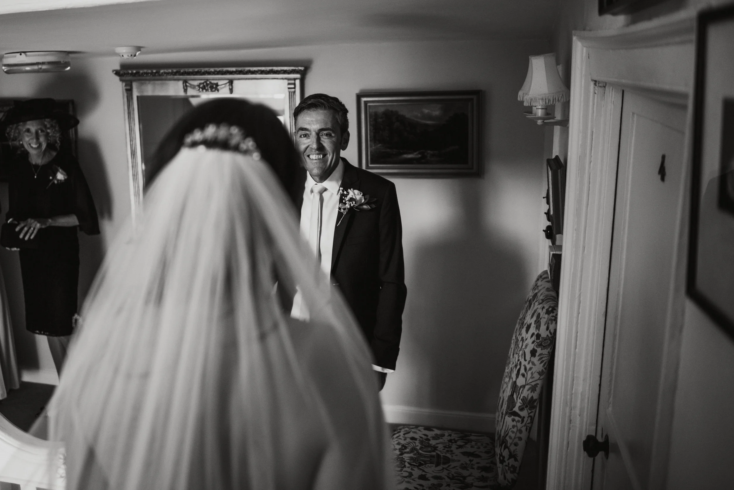 A bride in a veil and wedding dress looking into a mirror with an excited groom smiling at her. A woman in dark clothing and a wide-brimmed hat in the background smiling.