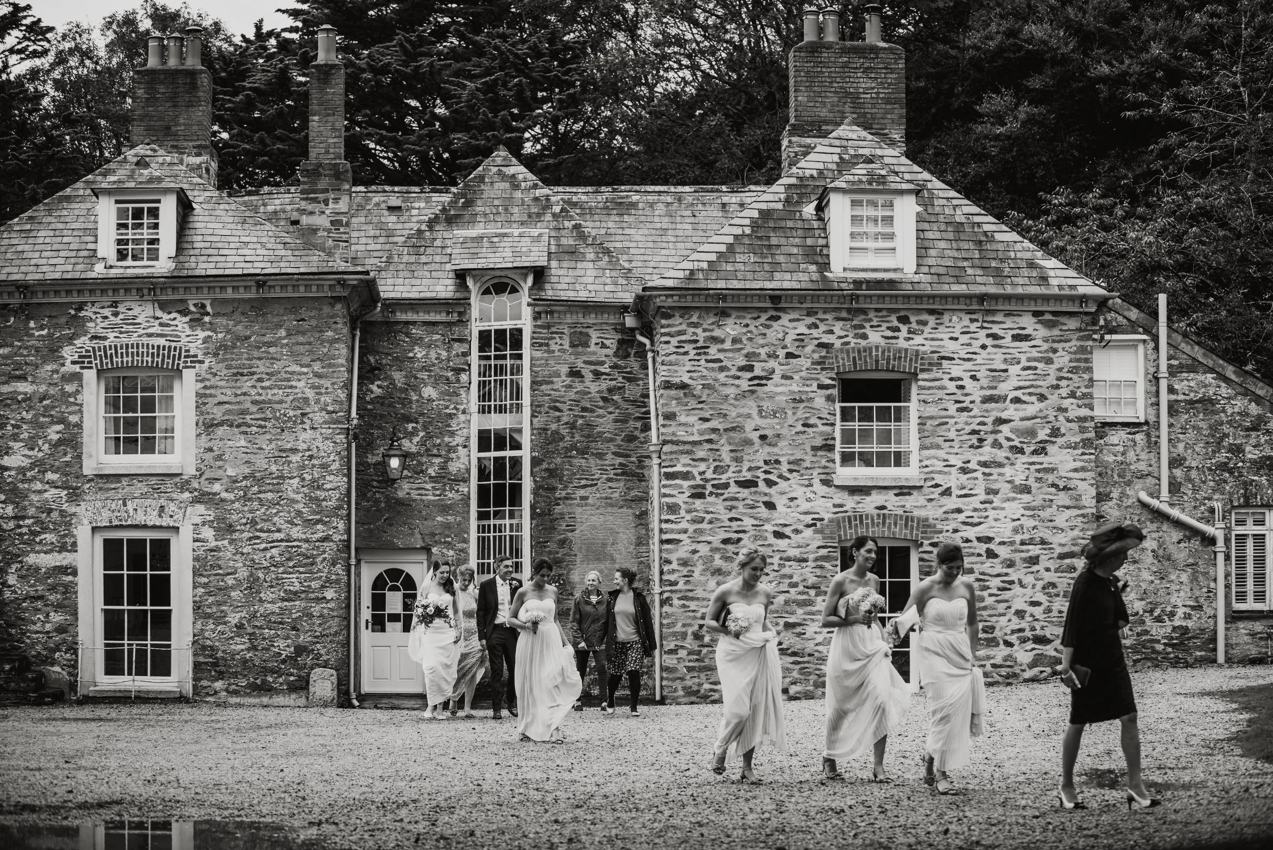 Black and white photo of a group of women in wedding dresses and a man in a suit, walking outside a stone house with multiple chimneys and windows, with trees in the background.