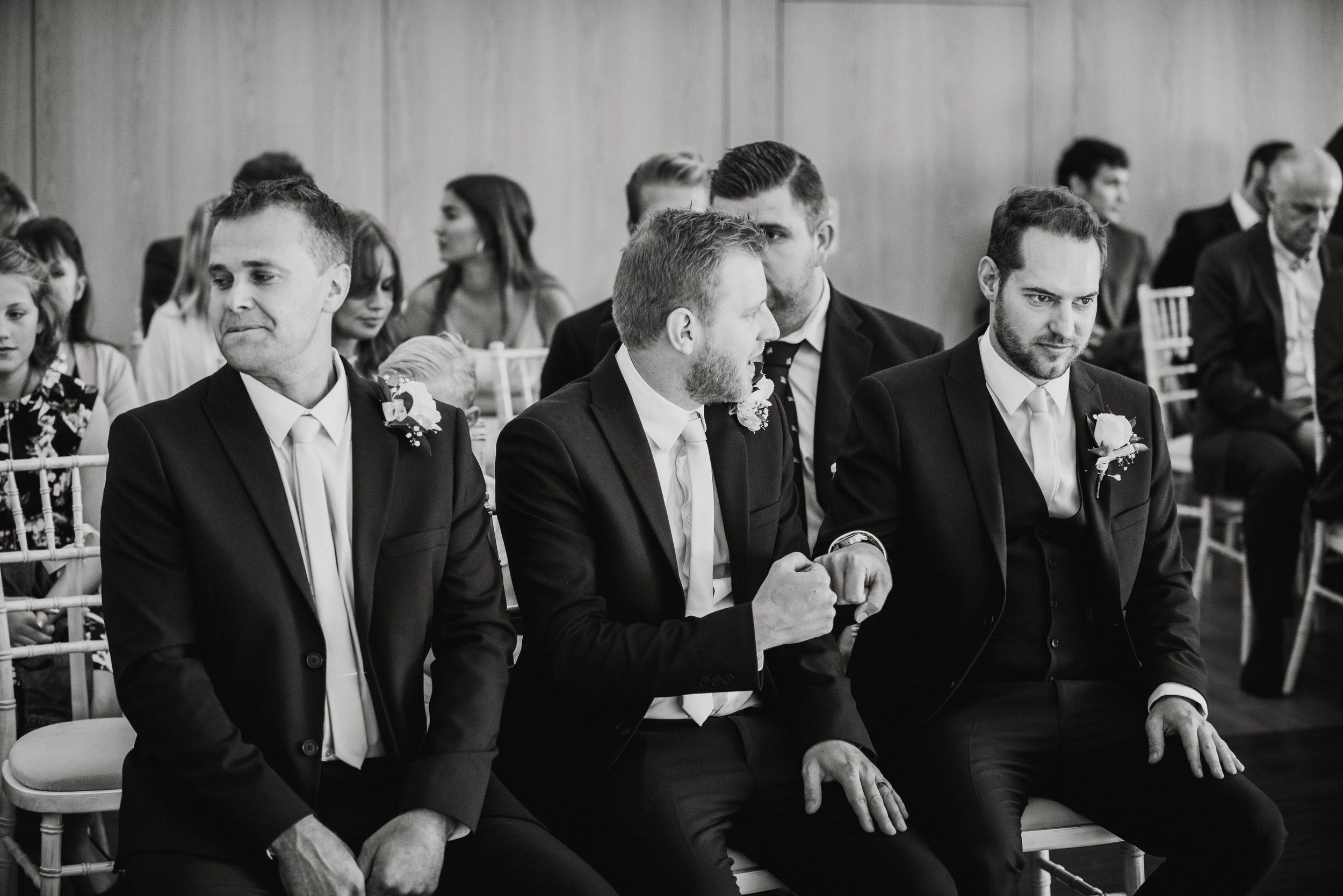 Black-and-white photograph of a group of men in suits sitting in chairs at a formal event, likely a wedding, with one man in the center talking to a man to his right.