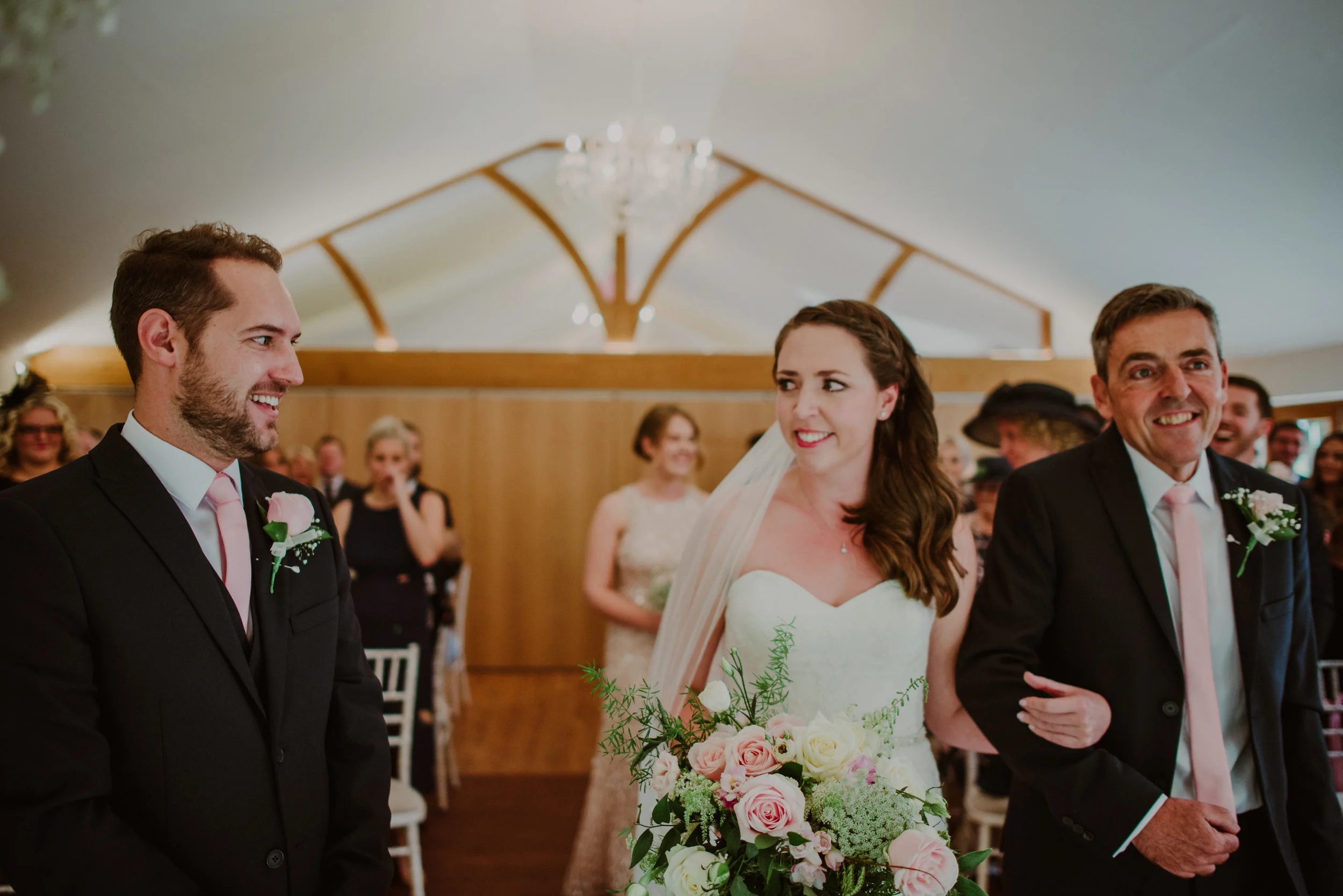 Bride walking down the aisle with her father at a wedding ceremony, surrounded by guests indoors.