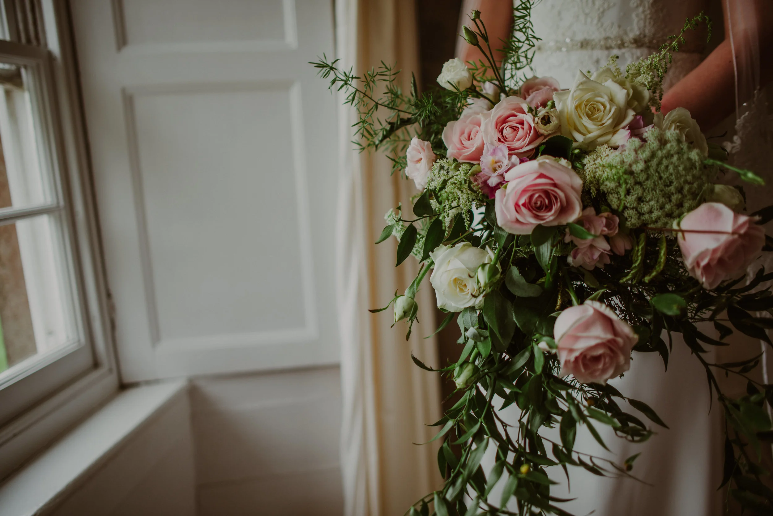 A bouquet of pink and white roses along with greenery, held indoors near a window with cream curtains.