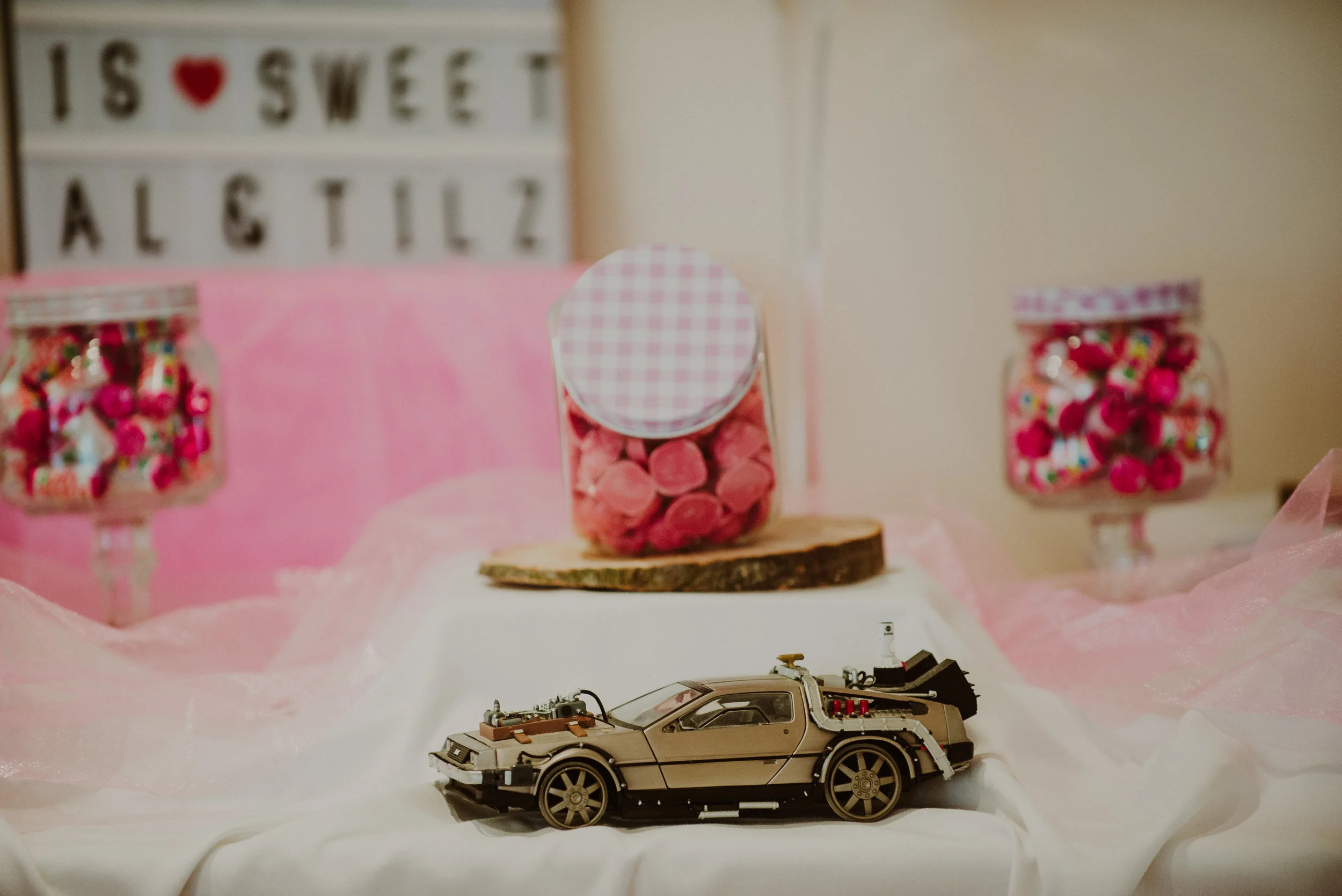 A miniature replica of the DeLorean car from 'Back to the Future' placed on a table with pink and white decorations, including jars of pink candy and a sign in the background reading 'I love sweet'.
