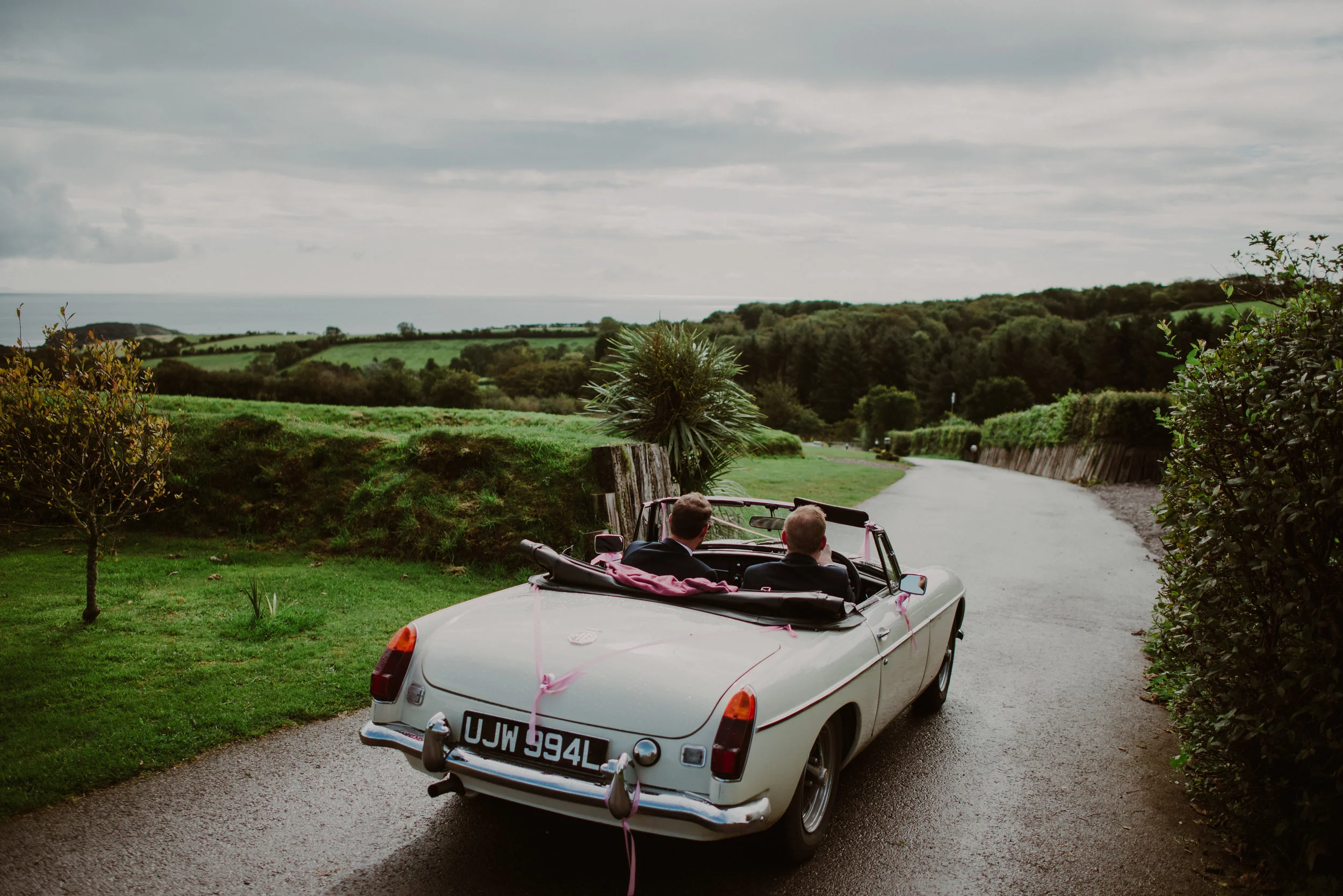A vintage white convertible car with a pink ribbon, carrying two men dressed in formal suits, driving on a winding rural road surrounded by green fields and trees under a cloudy sky.