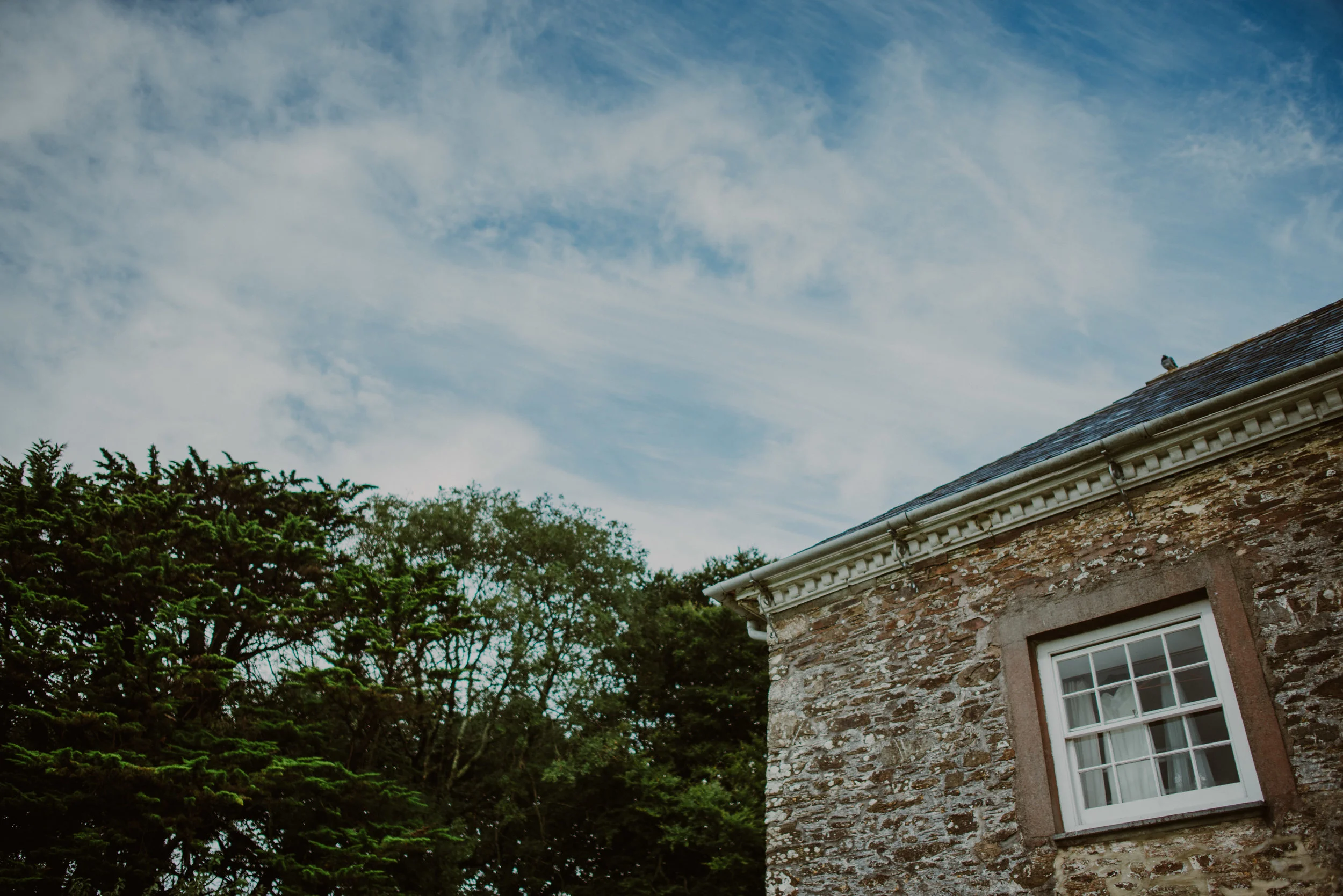 A corner of an old stone building with a window, surrounded by green trees and a cloudy sky.