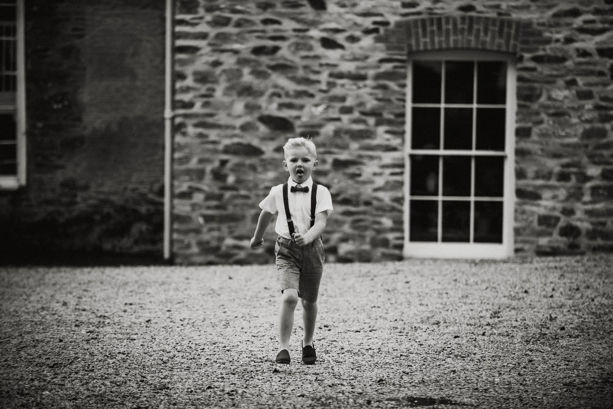 A young boy running towards the camera with an open mouth, dressed in a white shirt, bow tie, suspenders, and shorts, outside a stone building with a window and door.