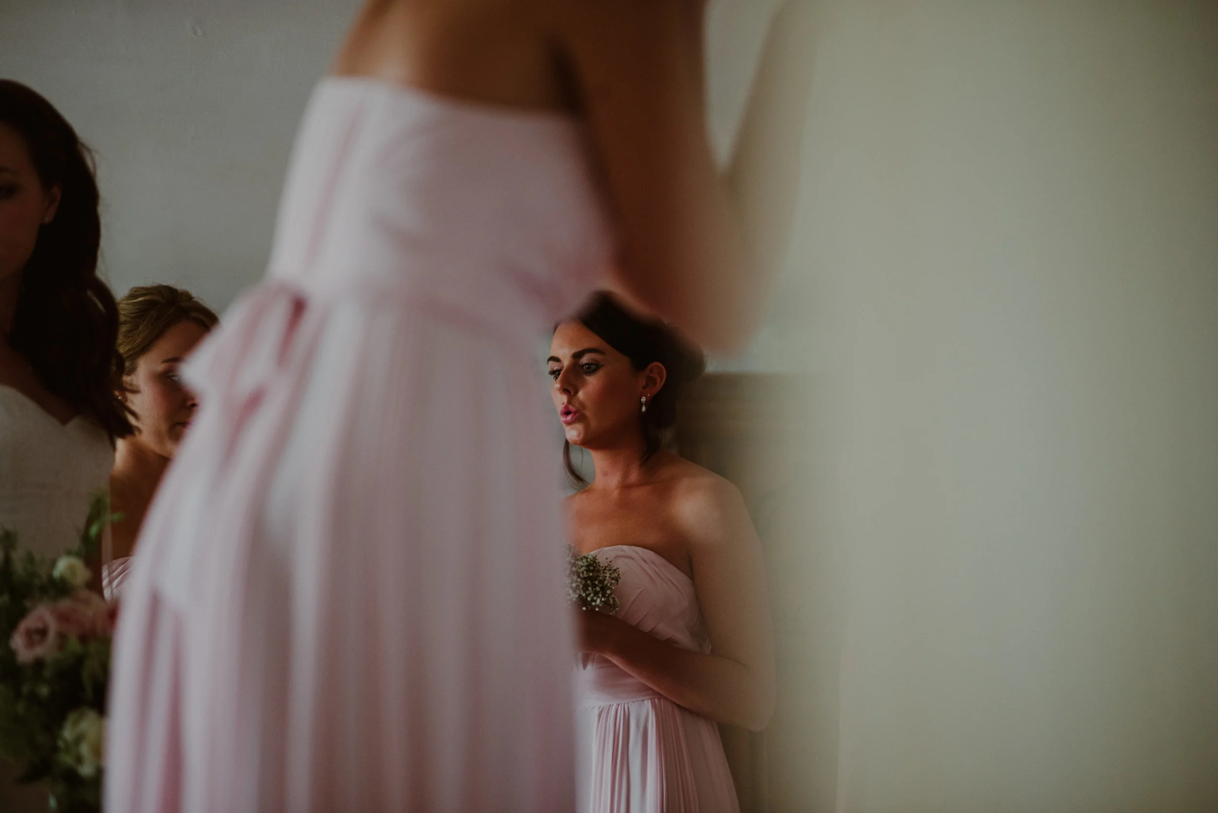 A woman in a pink dress is standing against a wall, holding a small bouquet, with other women in similar dresses partially visible around her.