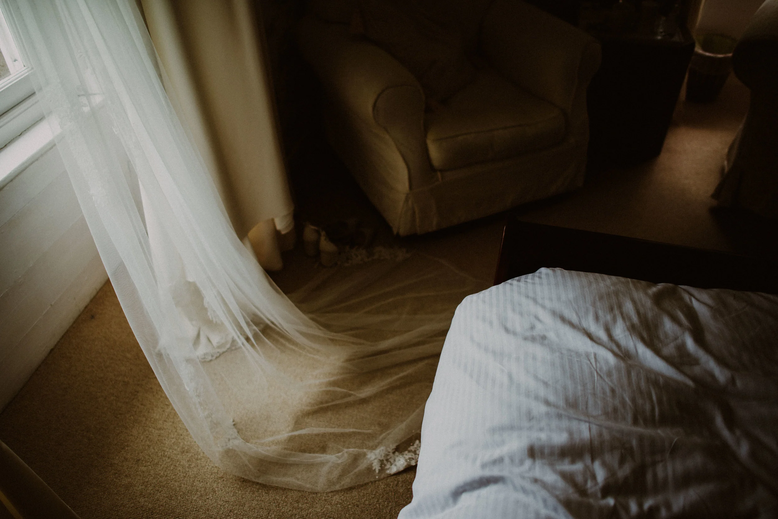 A corner of a bedroom with a bed in the foreground, a sheer curtain hanging from a window, a beige armchair and another armchair in the background, and a cat partly hidden under the armchair.