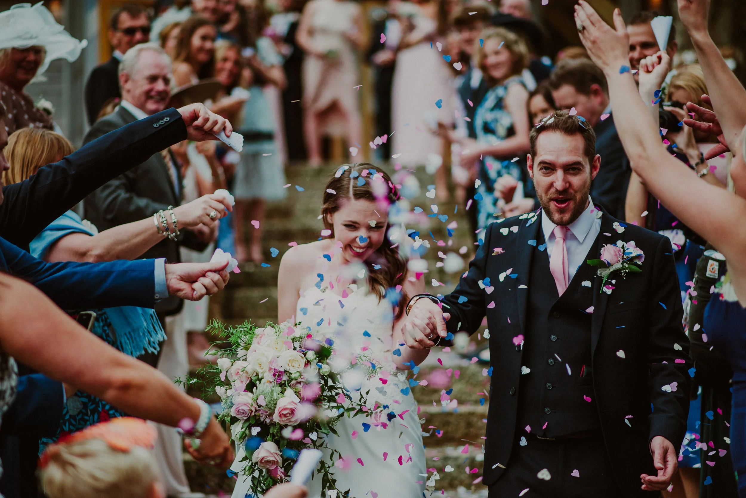 A newlywed couple holding hands and smiling as friends and family toss colorful confetti during their outdoor wedding celebration.