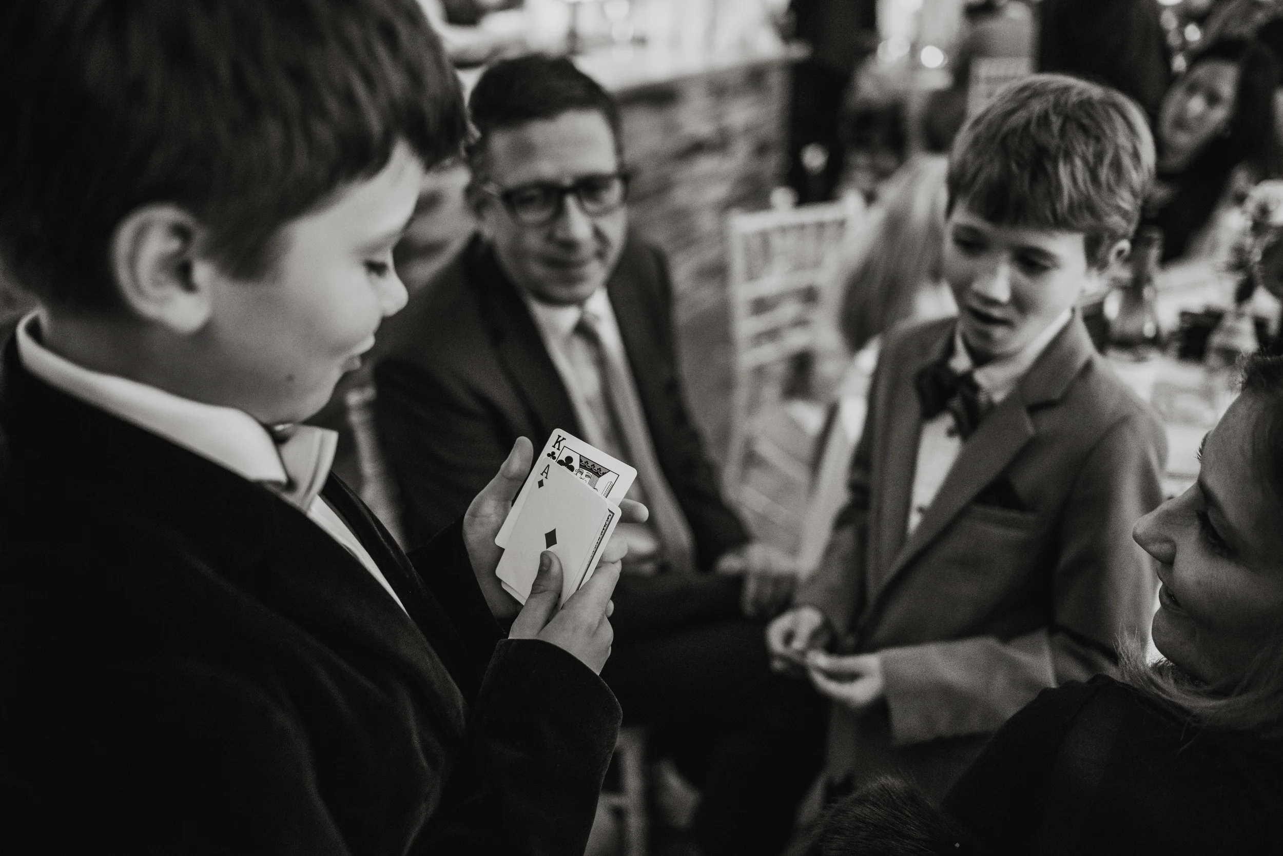 A young man in a tuxedo holding playing cards, with others around him, at a social gathering.