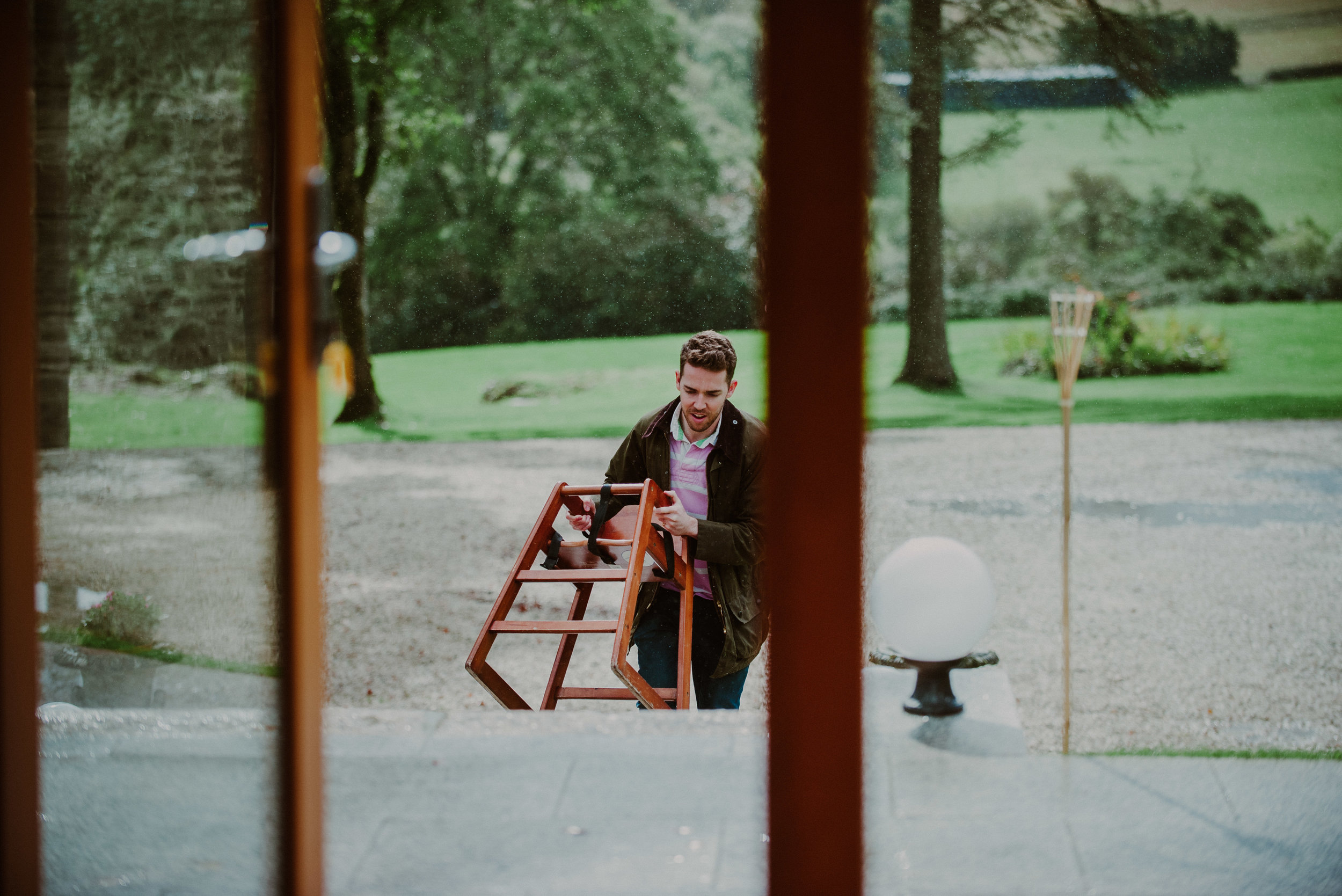A man seen through a window, holding a wooden chair outdoors on a gravel surface, with a green landscape and trees in the background.
