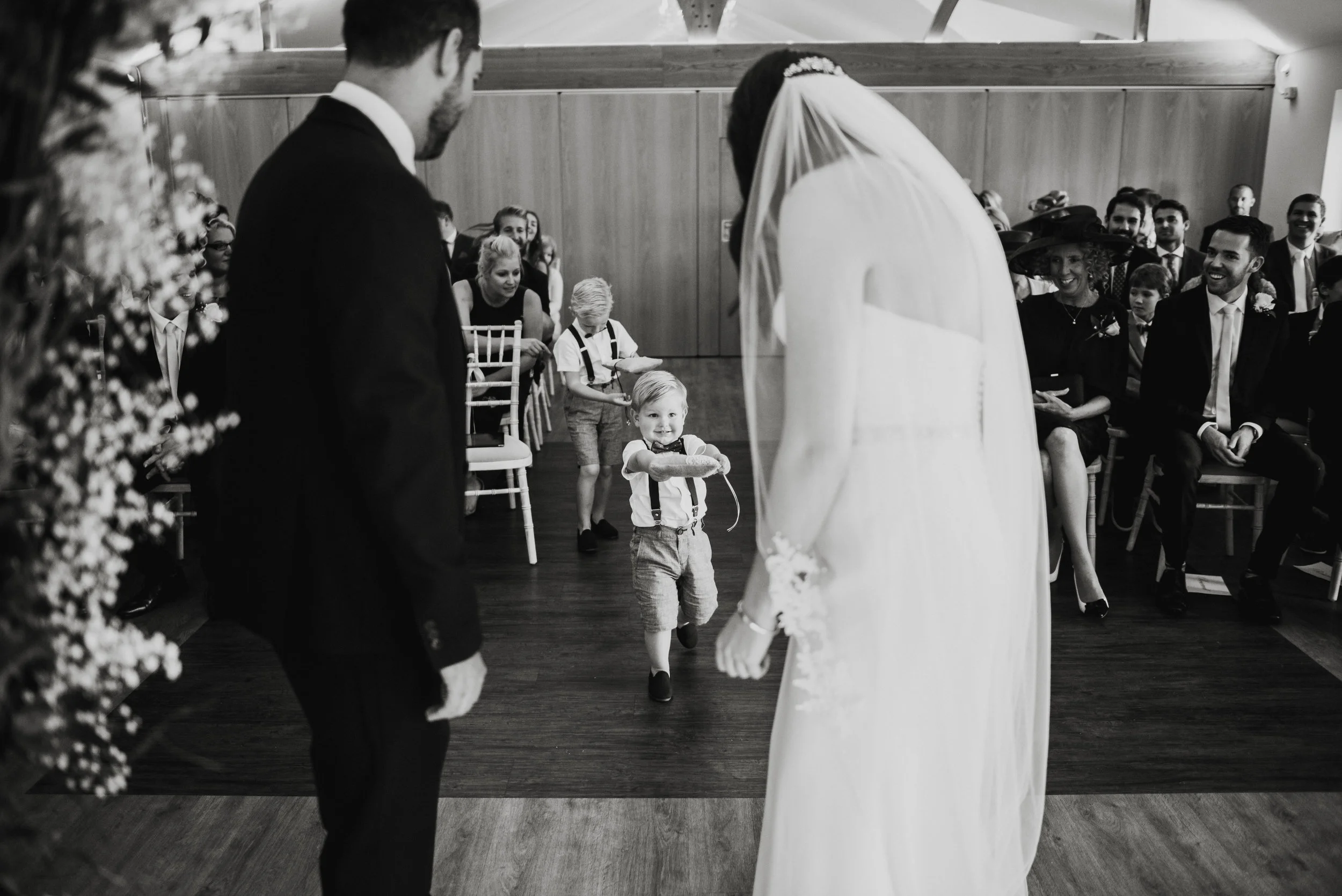 A wedding ceremony taking place indoors with the bride and groom at the front. The bride wears a white dress and veil; the groom in a dark suit. In the background, guests are seated and smiling, and two children are walking towards the couple, one of