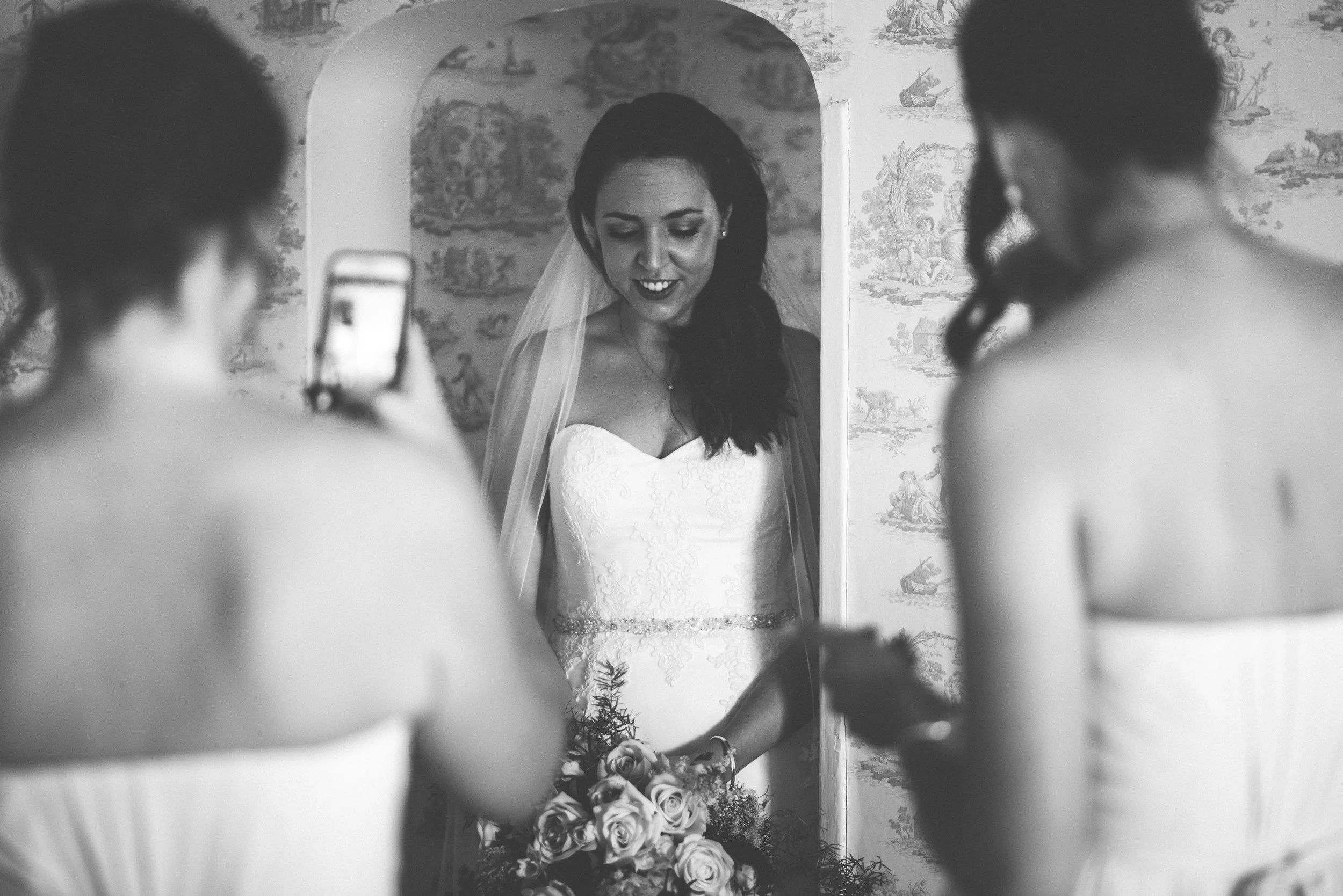 A bride in a wedding dress looking at her reflection in a mirror, with two women taking photos of her.