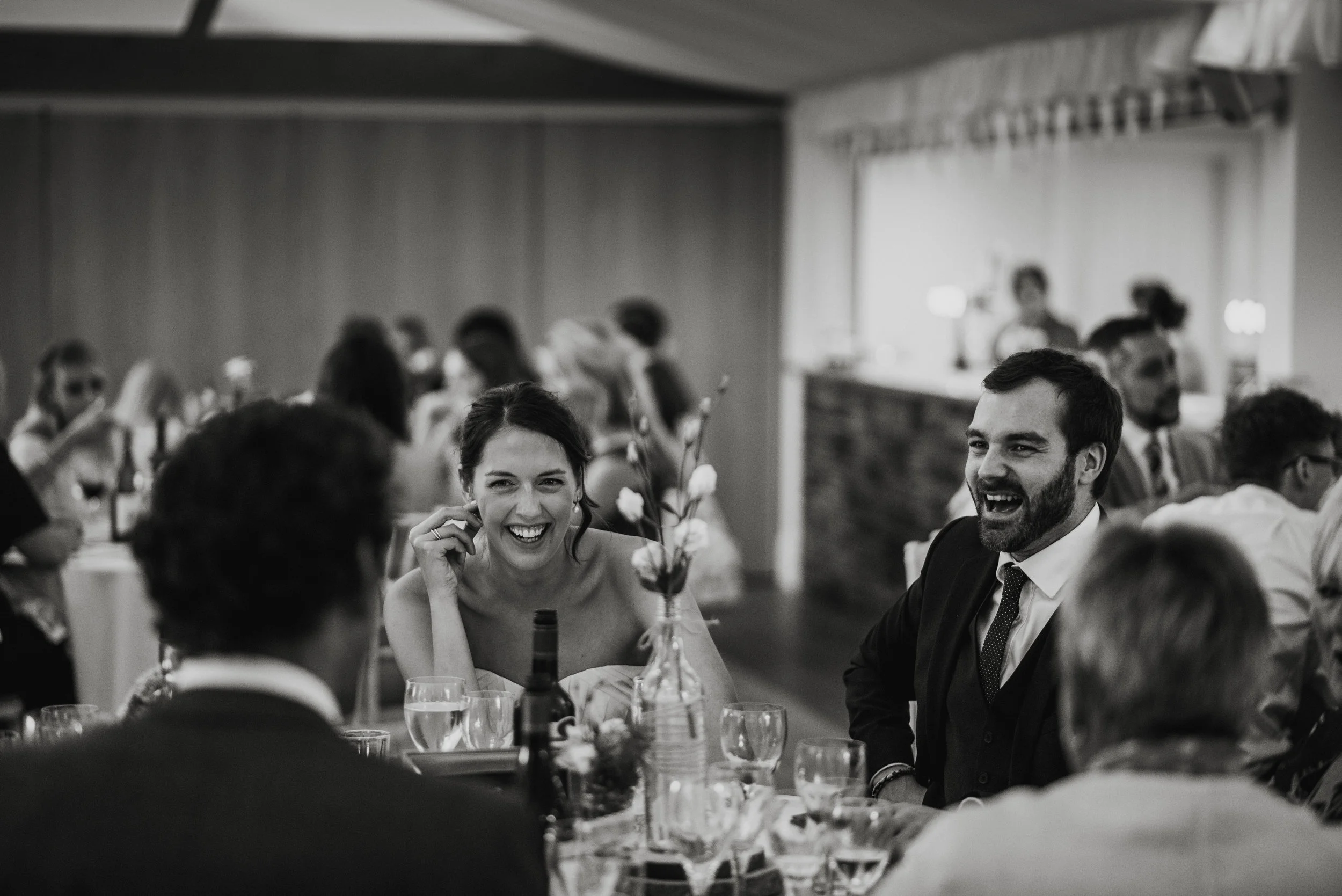Black and white photo of a group of people at a formal event or wedding reception, with a woman and man in the foreground smiling and laughing, seated at a decorated table.