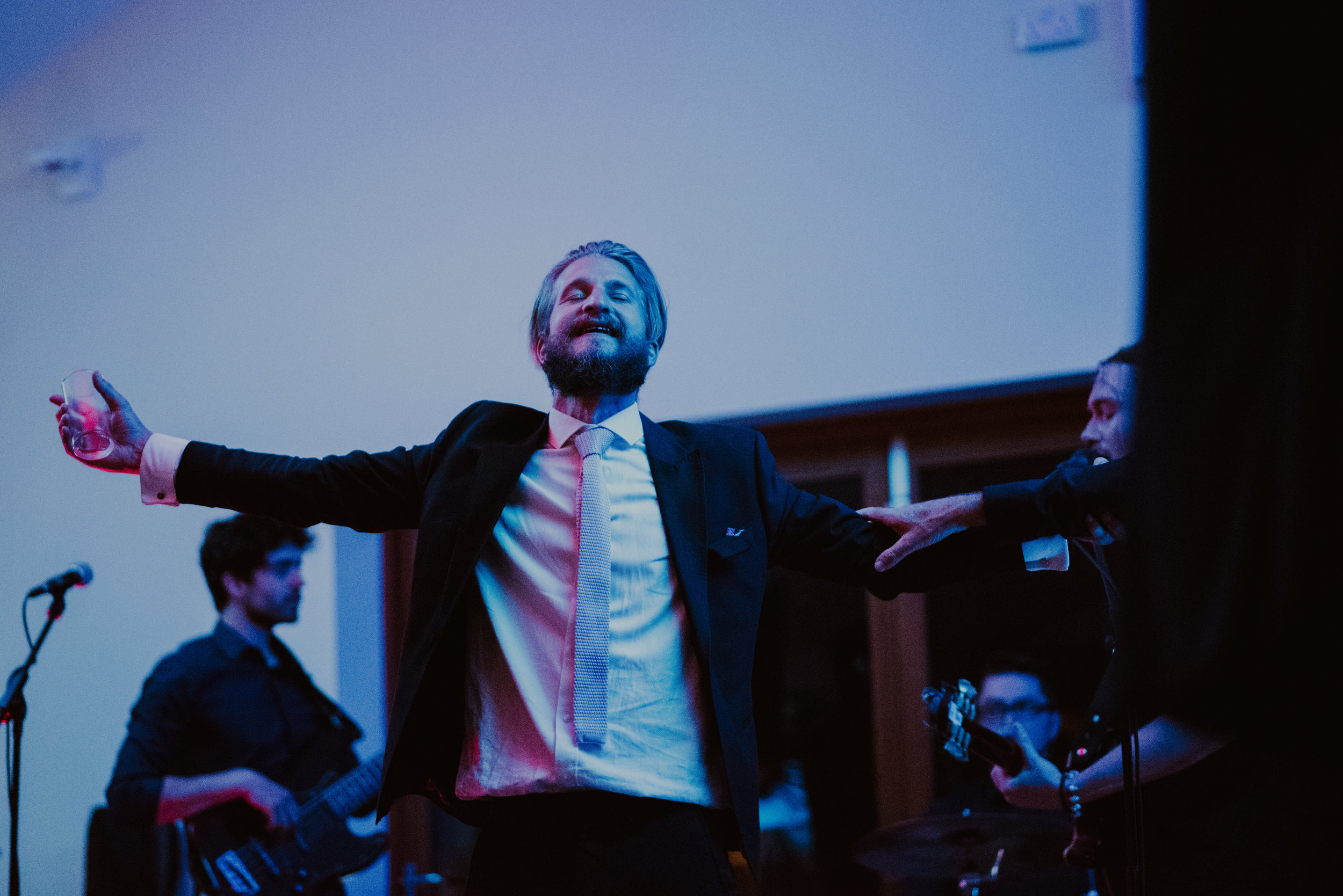 A man in a suit with open arms and a glass in hand, enjoying a moment during a musical performance with band members playing guitars in the background.