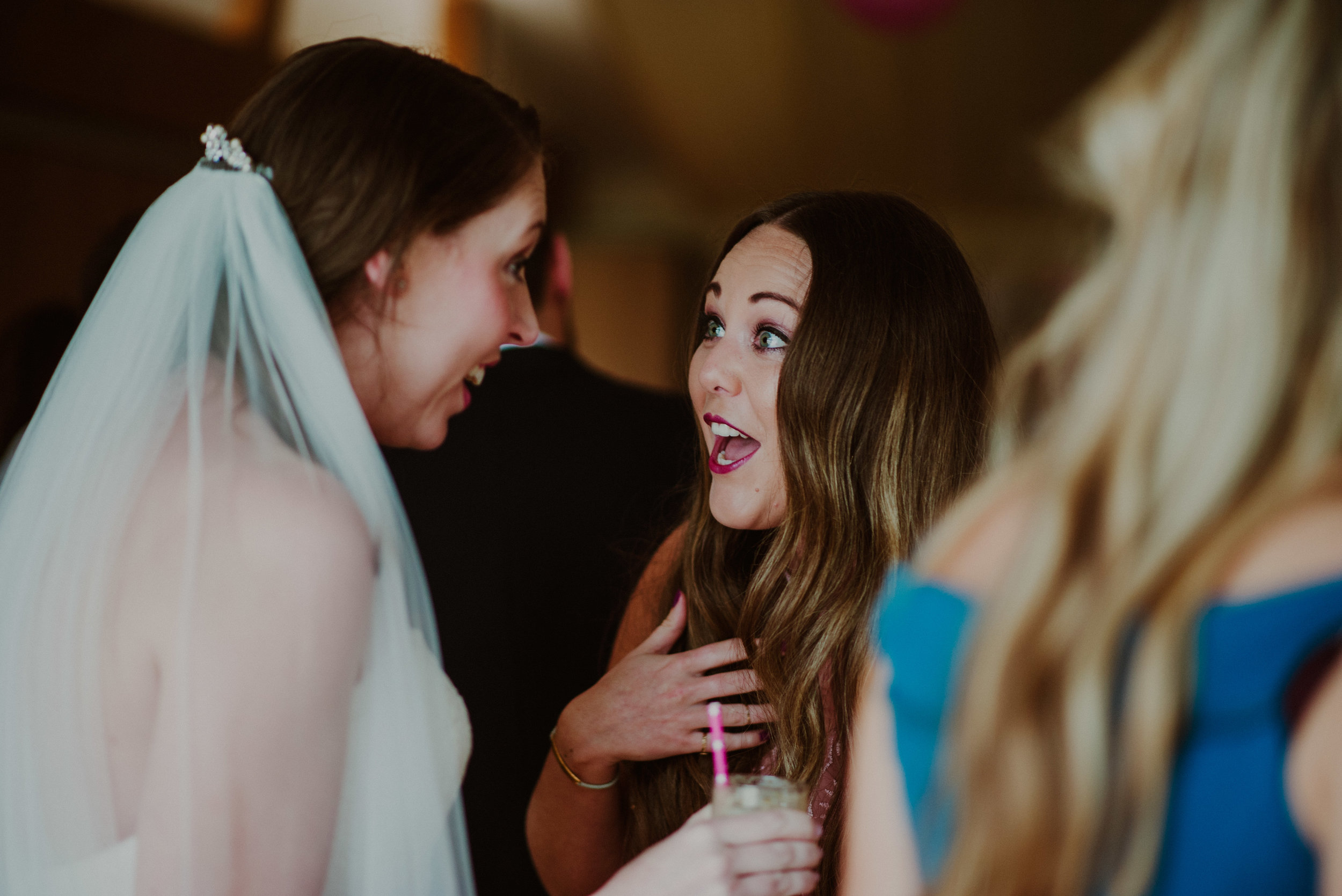 Two women at a wedding reception, facing each other and reacting with surprise and excitement, one in a wedding dress with a veil, and the other holding a drink with a pink straw.