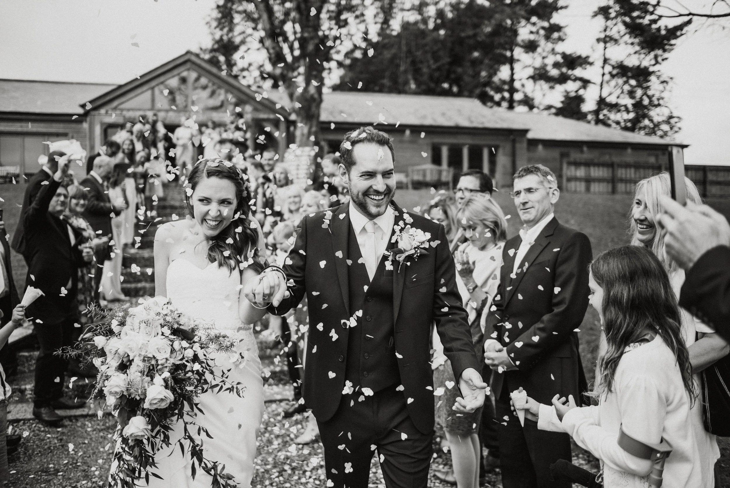 A newlywed couple walking hand in hand through a crowd as guests throw confetti during an outdoor wedding celebration.