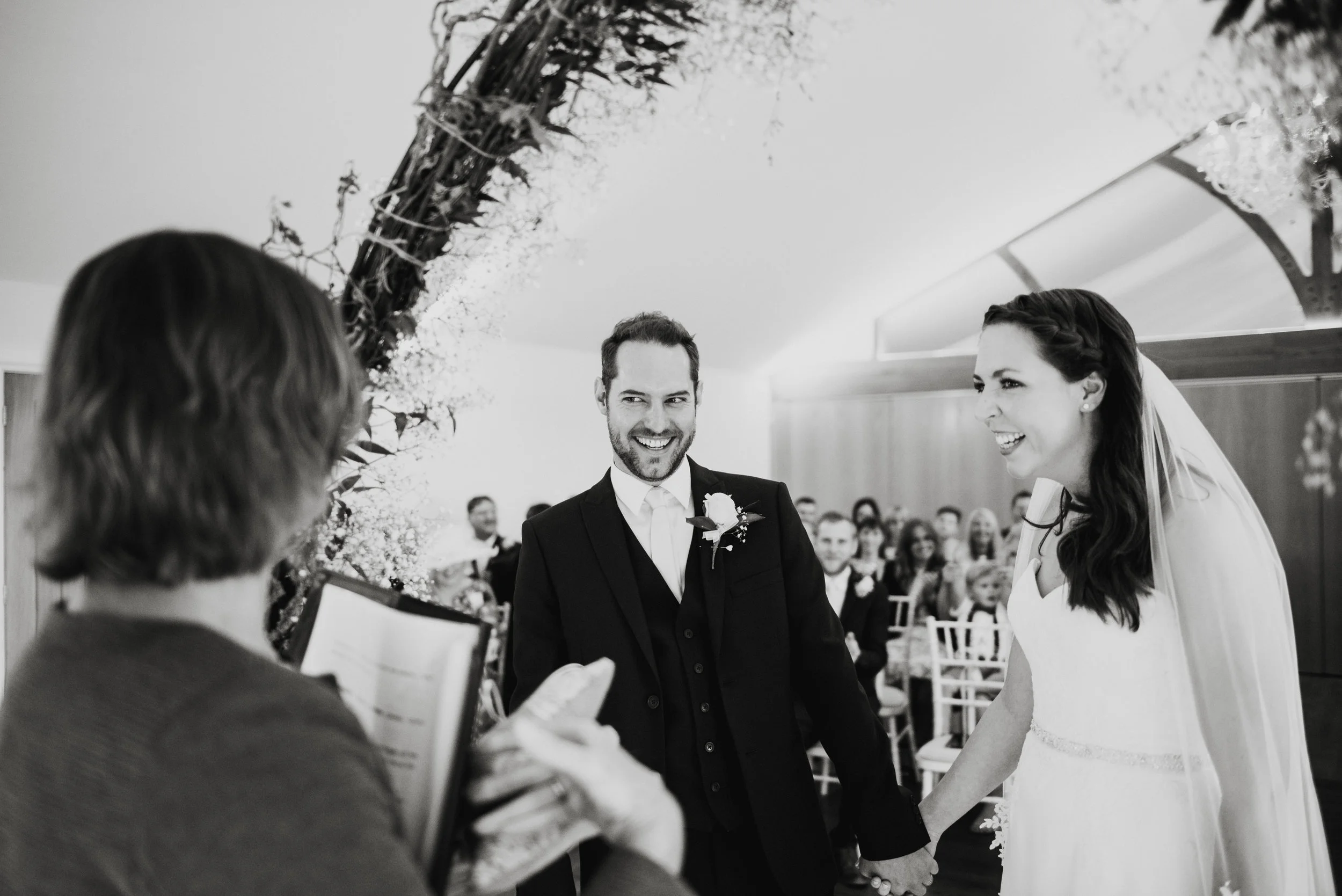 A wedding ceremony with a couple holding hands, smiling at each other, surrounded by seated guests, with a floral arch above them.