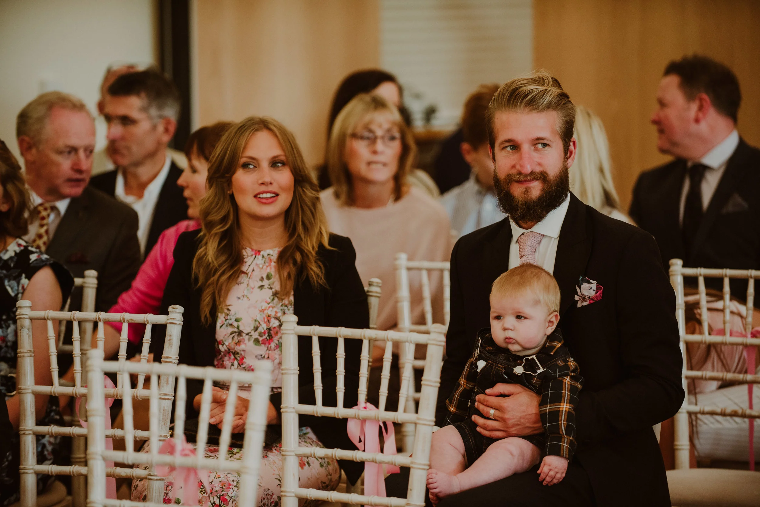 People attending a wedding ceremony, with a man holding a baby in the foreground, and women and men in formal attire seated behind them.
