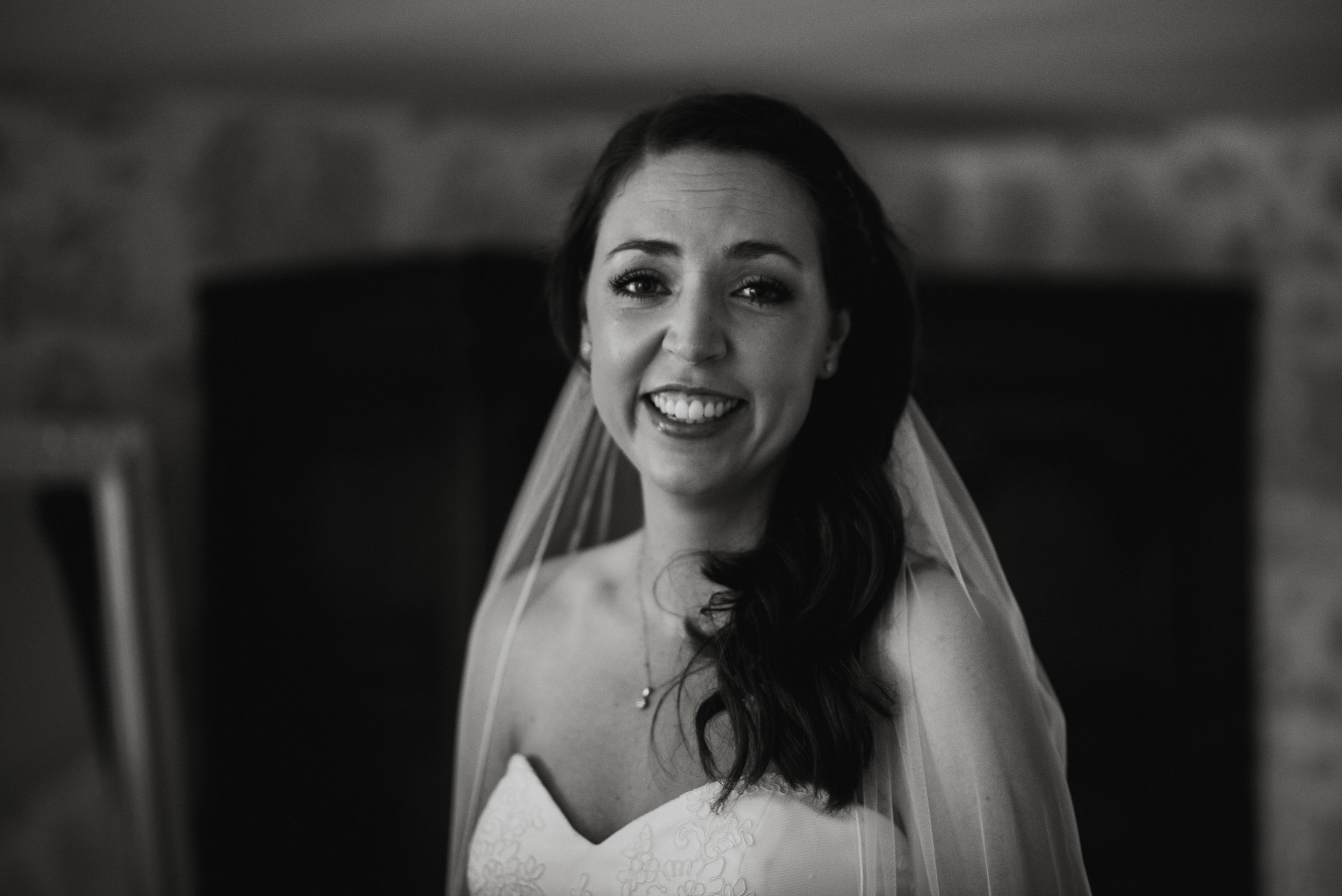 A smiling woman in a wedding dress wearing a veil, looking at the camera, in black and white.