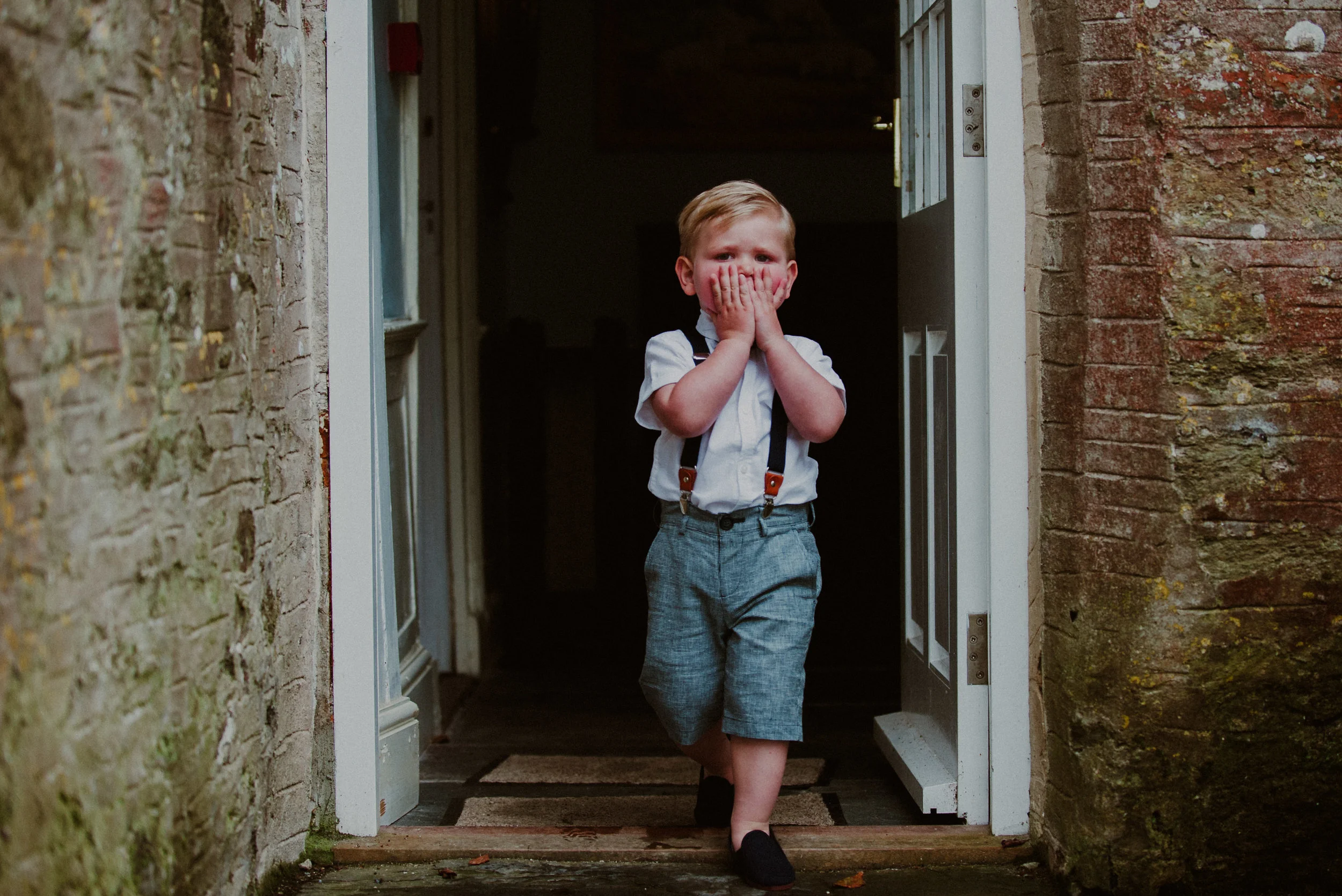 A young boy wearing a white shirt, gray shorts with suspenders, and black shoes, standing in a doorway with his hands covering his mouth and nose, looking surprised or excited.