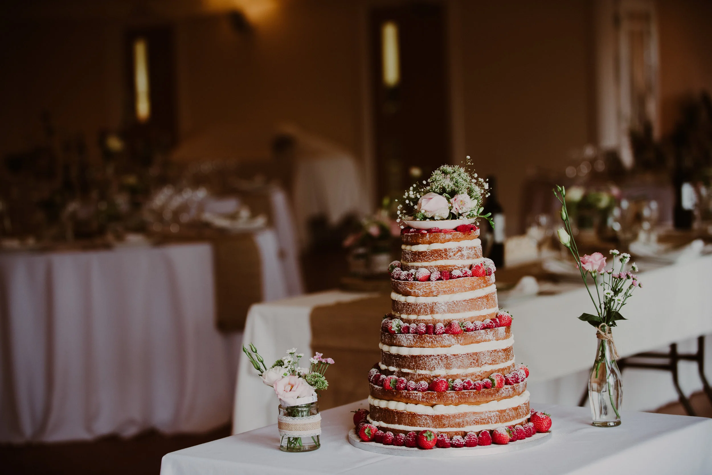 A tiered naked wedding cake decorated with fresh strawberries and topped with flowers, flanked by small flower vases on a table.