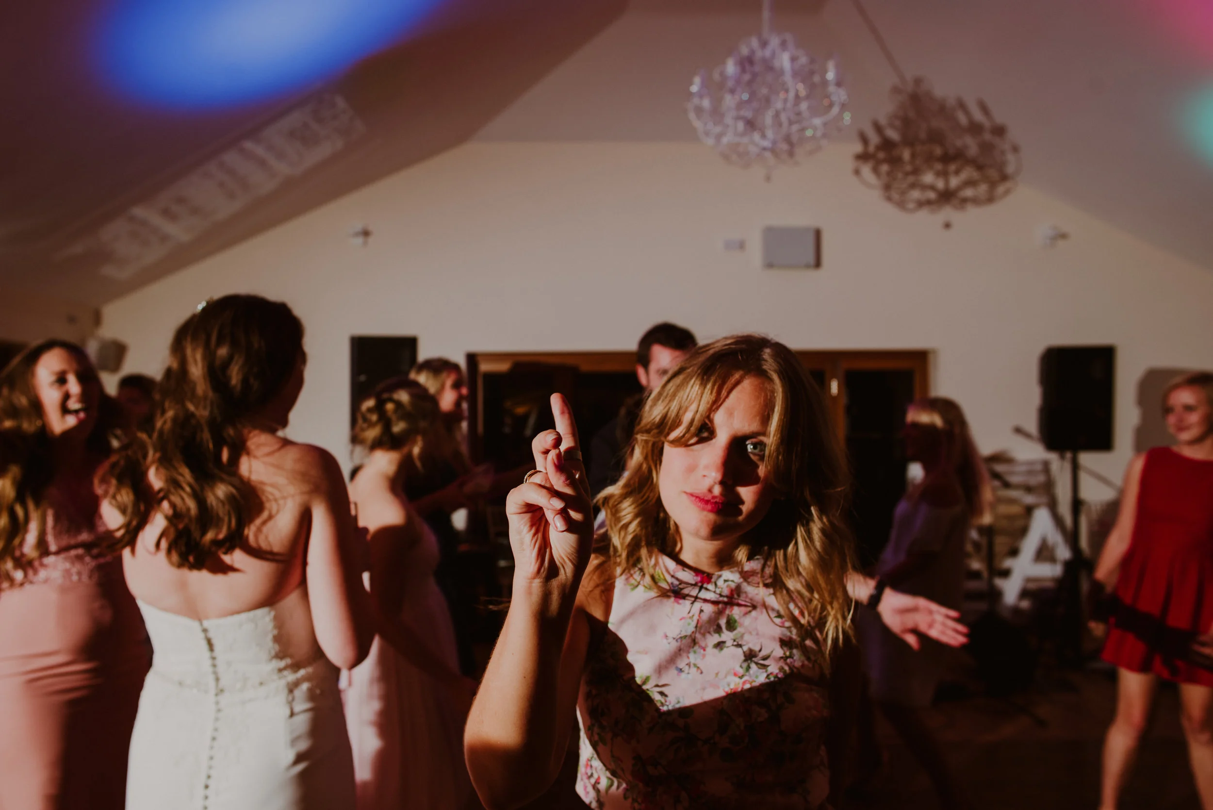 A woman in a floral dress dancing and pointing her finger at a party or wedding reception, surrounded by other women in dresses, with a chandelier and sound equipment in the background.