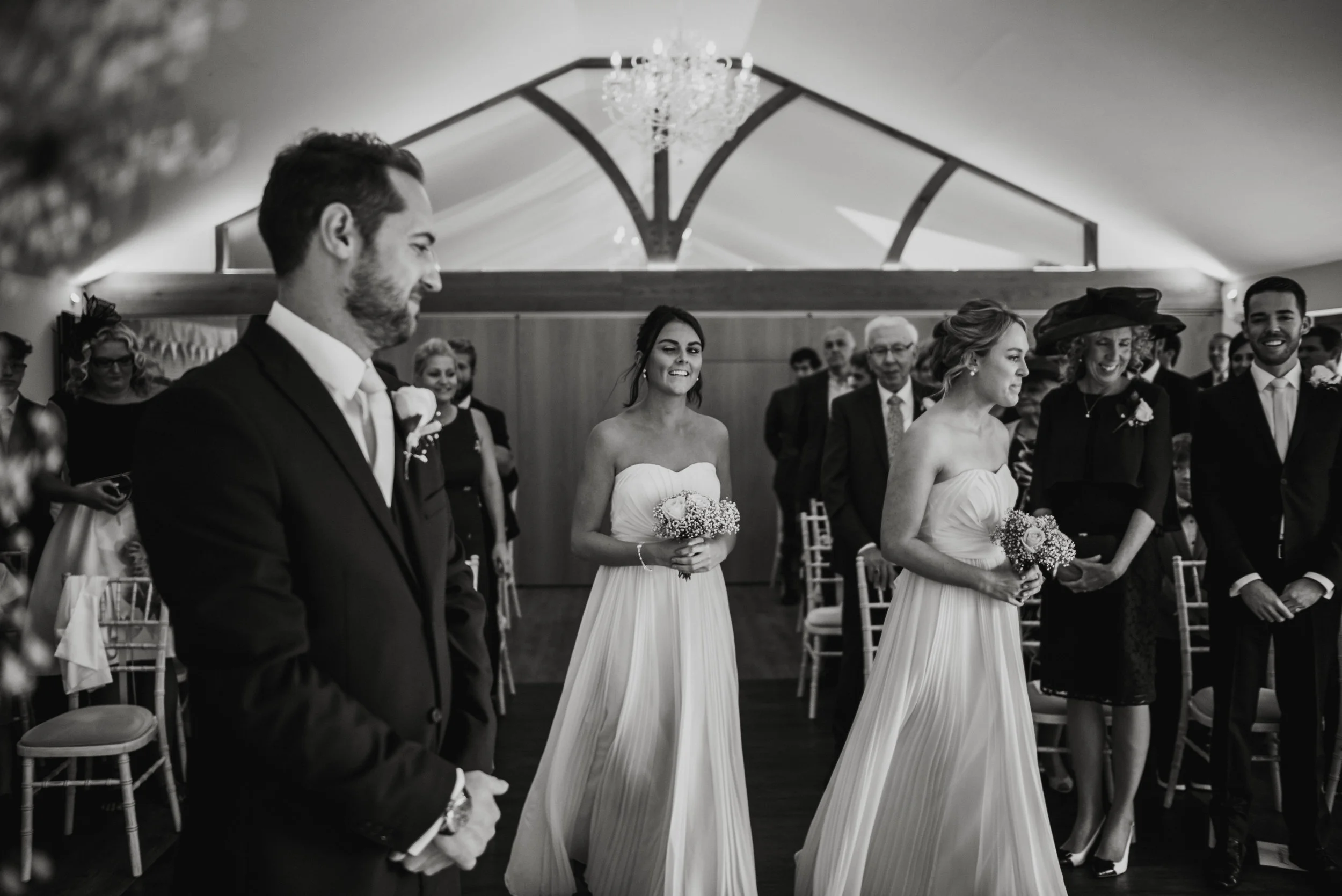 A black-and-white photo of a wedding ceremony with the bride, groomsmen, bridesmaids, and guests inside a decorated venue with a chandelier.
