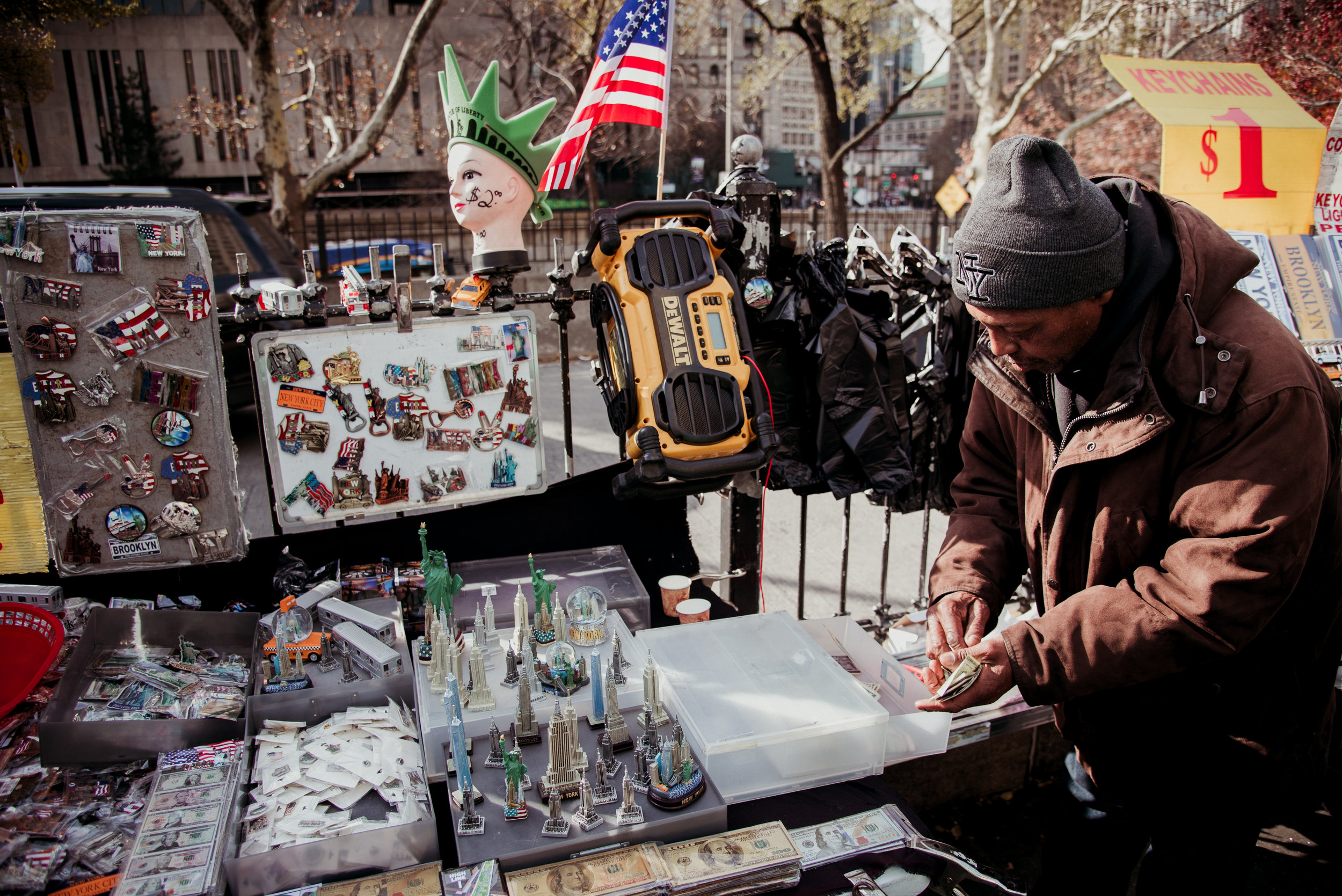A street vendor selling souvenirs and accessories, including keychains, pins, miniature models of landmarks, and cash, at an outdoor street market. The vendor is counting money, and there are various items on display, such as a Halloween mask, Americ