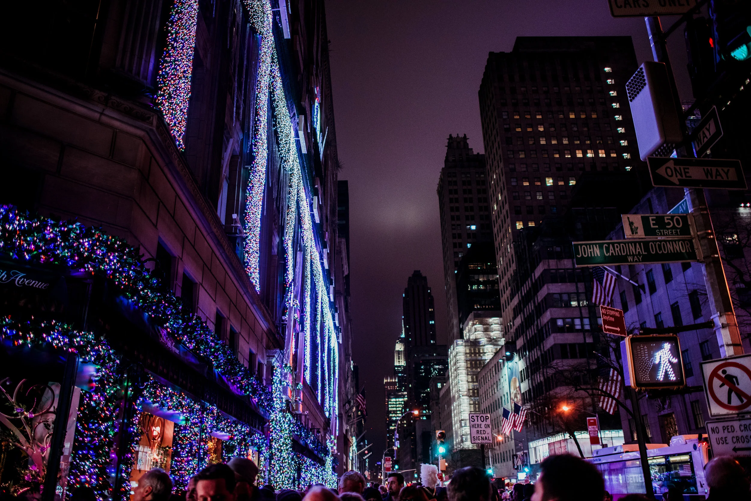 Nighttime street scene on a city block decorated with colorful Christmas lights and ornaments, with people walking and several tall buildings in the background.