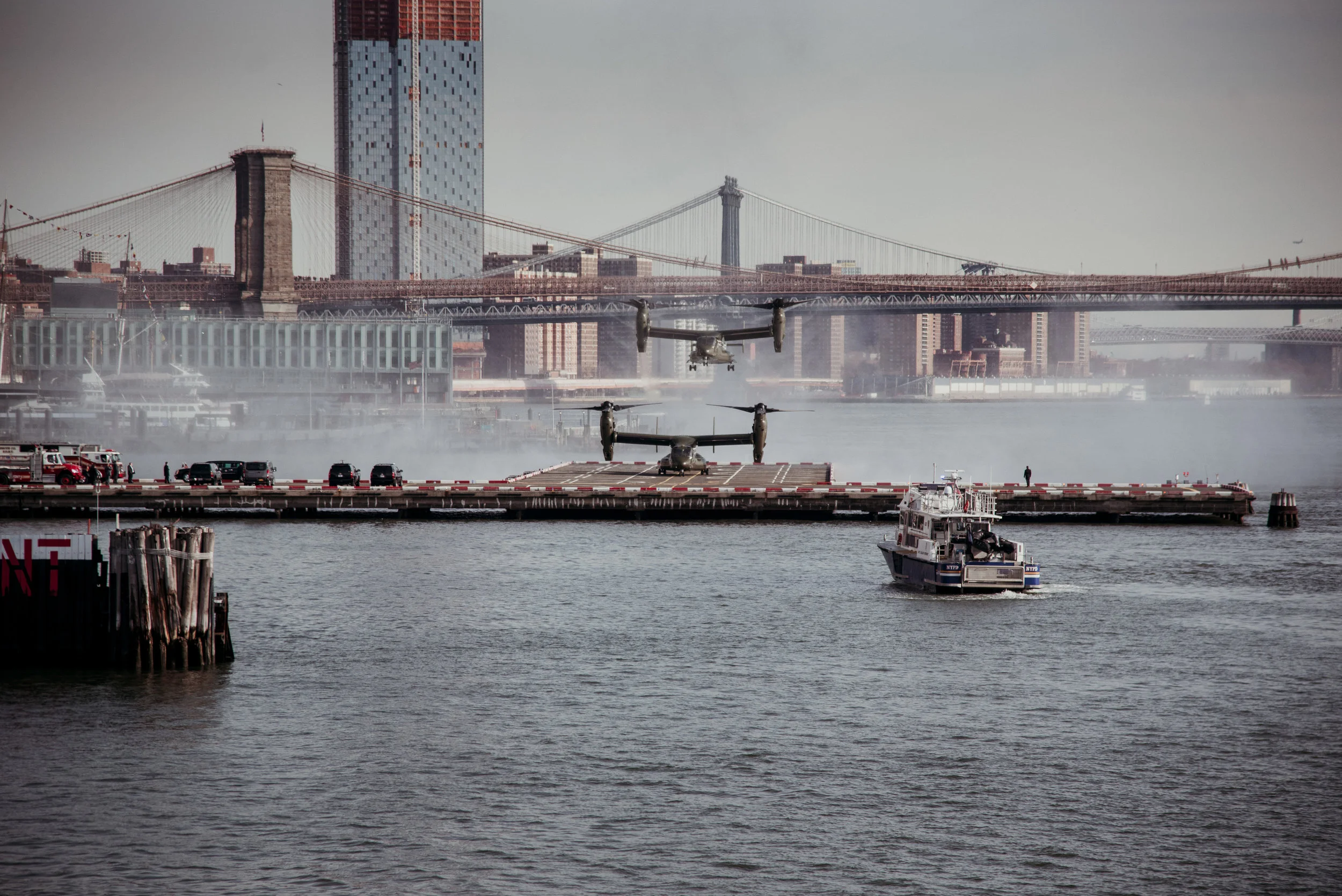 Two helicopters flying low over a pier with a boat in the water nearby, city skyline with bridges in the background.