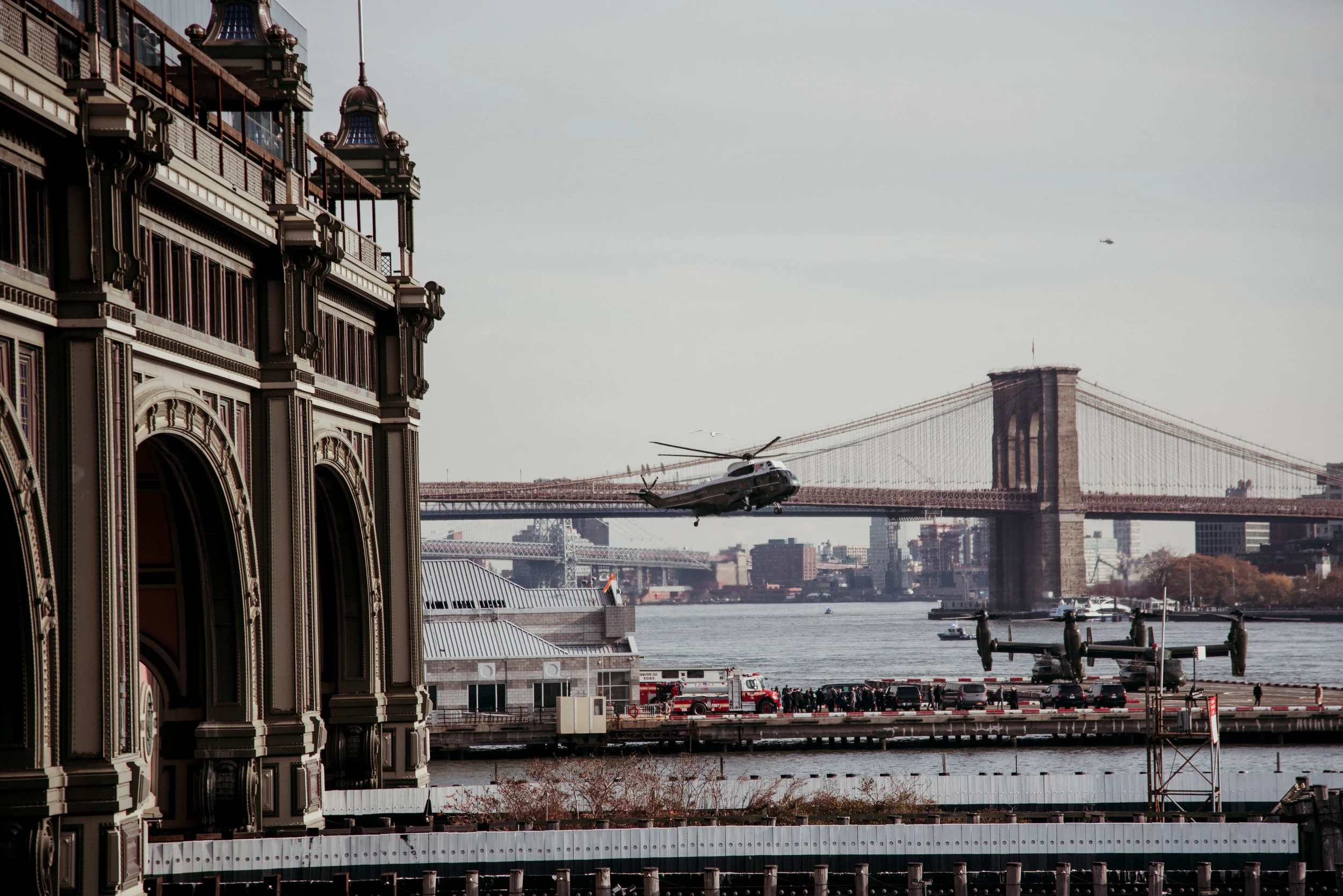 A helicopter flying over a waterfront with the Brooklyn Bridge in the background, near a historic building with ornate architecture, fire trucks, and military aircraft on display.