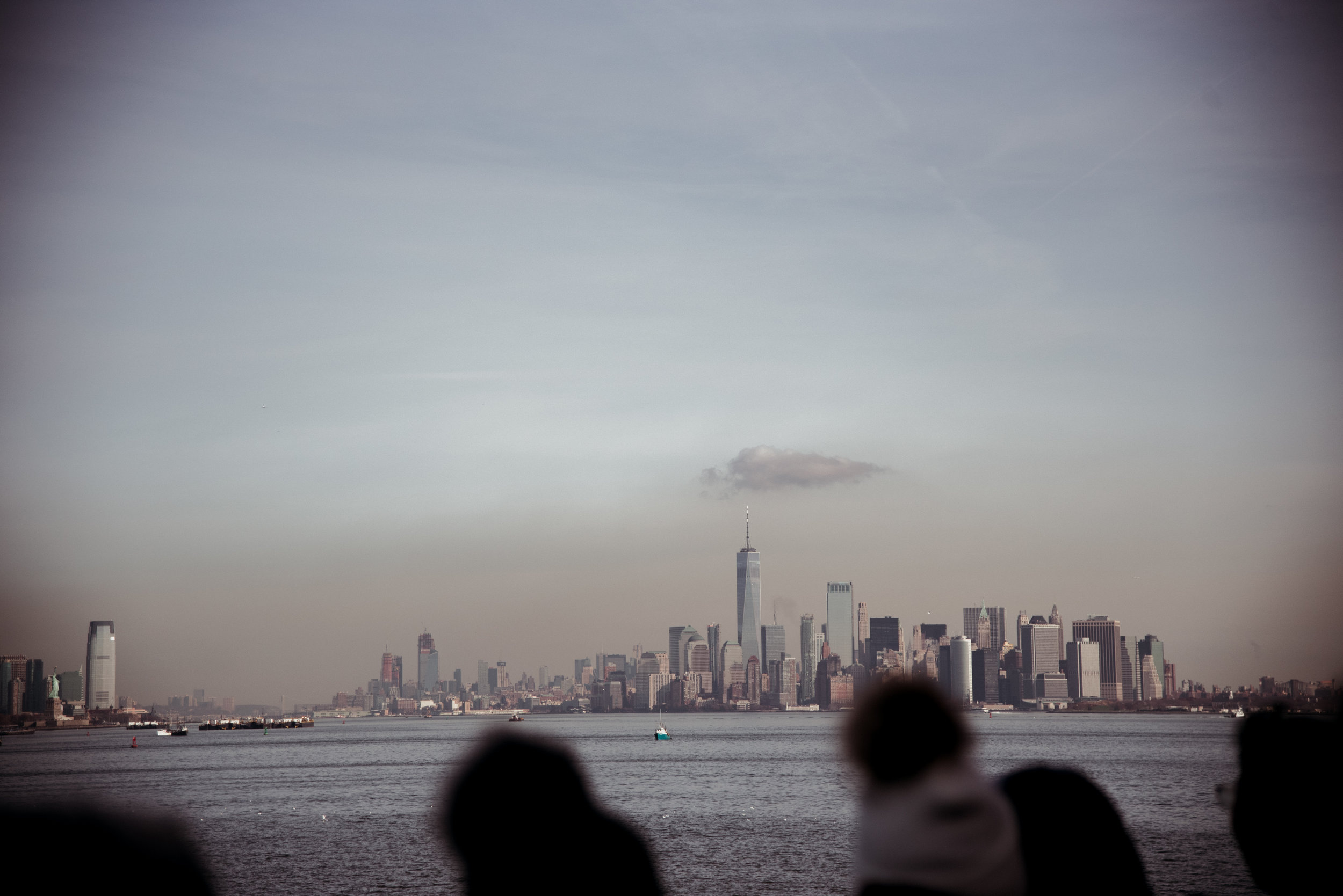 New York City skyline with tall buildings, including One World Trade Center, viewed across a body of water with silhouettes of people in the foreground, and a partly cloudy sky.