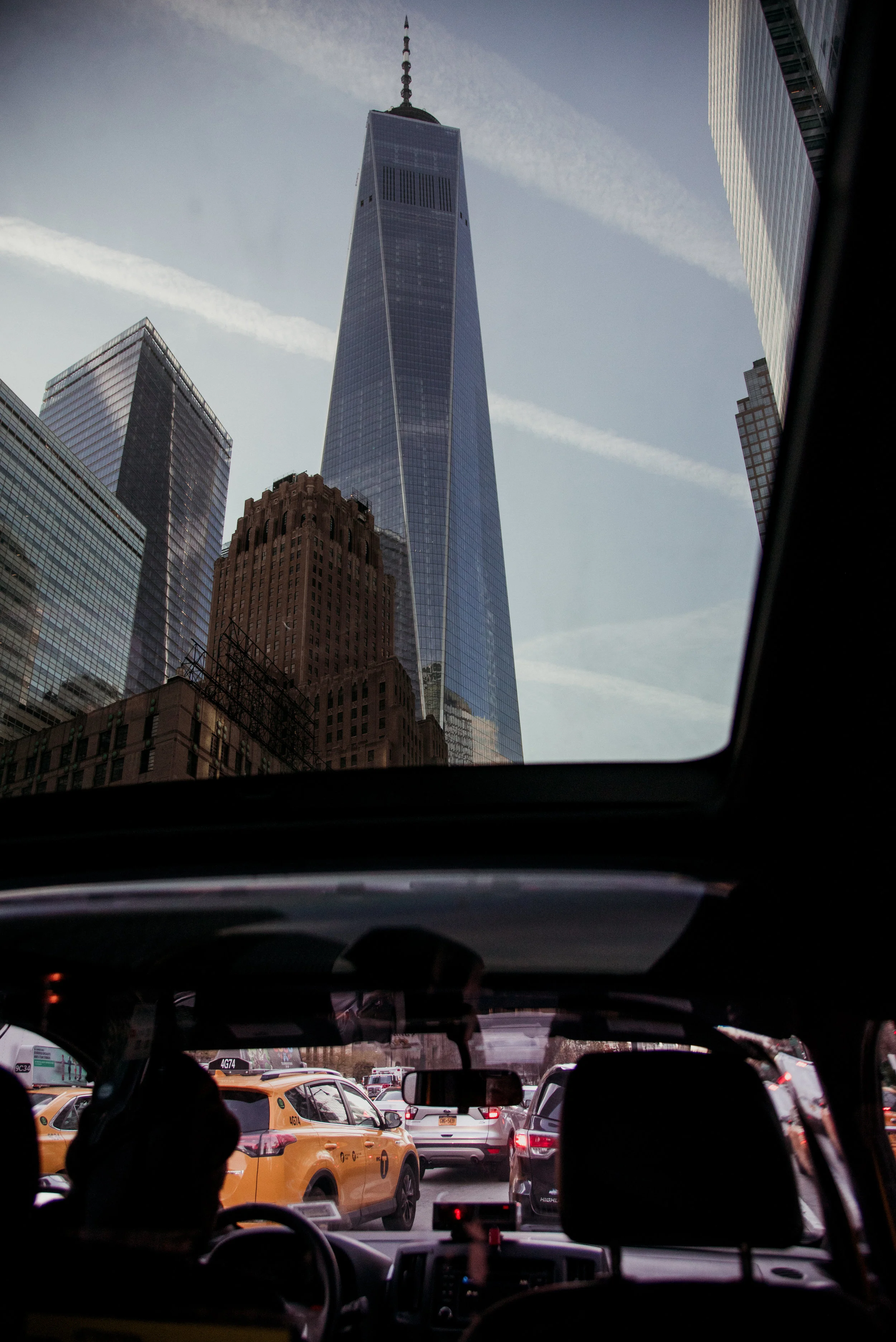 Cityscape viewed from inside a vehicle, with a view of One World Trade Center towering above other buildings, seen through the windshield, with yellow taxis and traffic below.