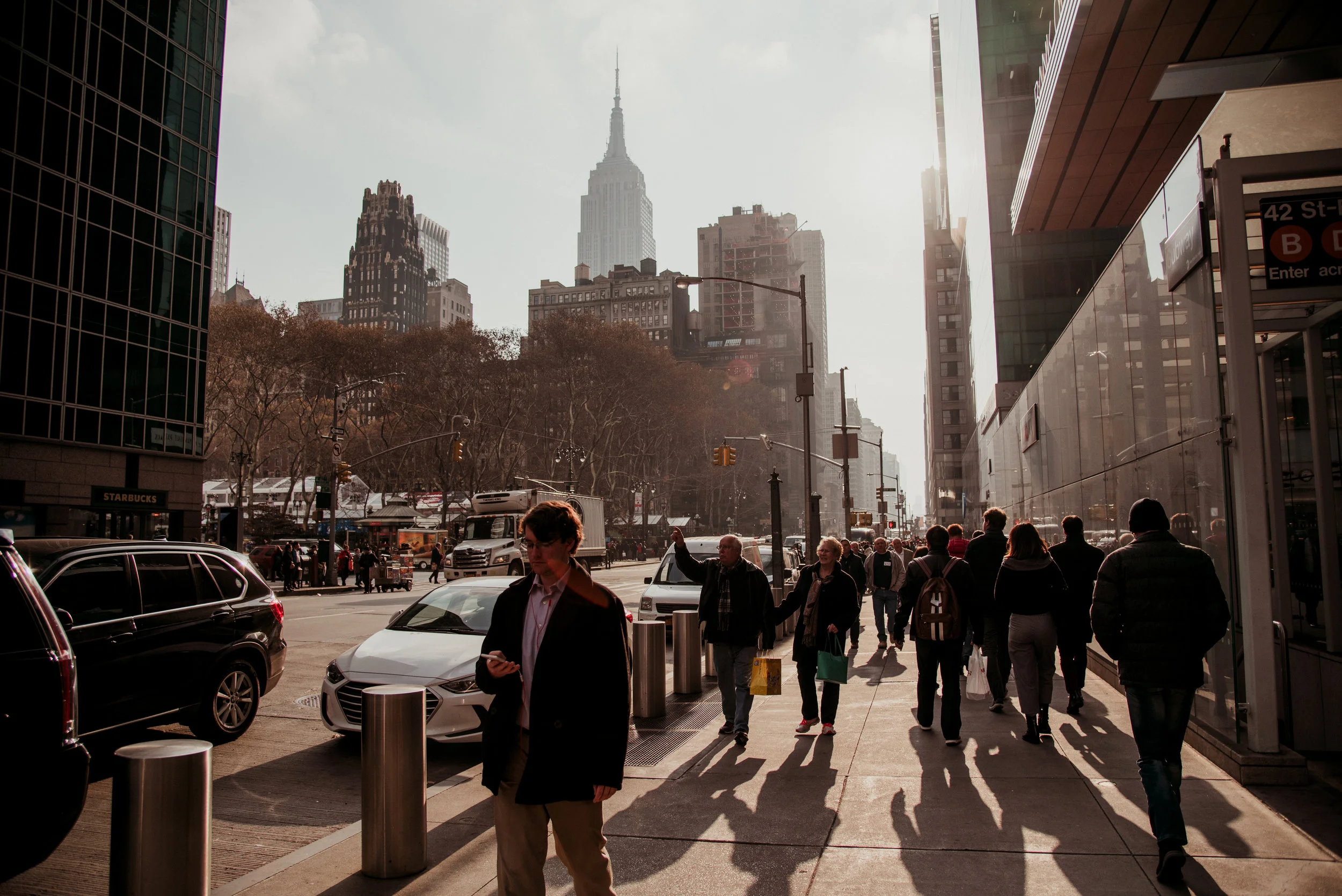 City street scene in New York with people walking on the sidewalk, tall buildings, and the Empire State Building in the background during late afternoon sun.