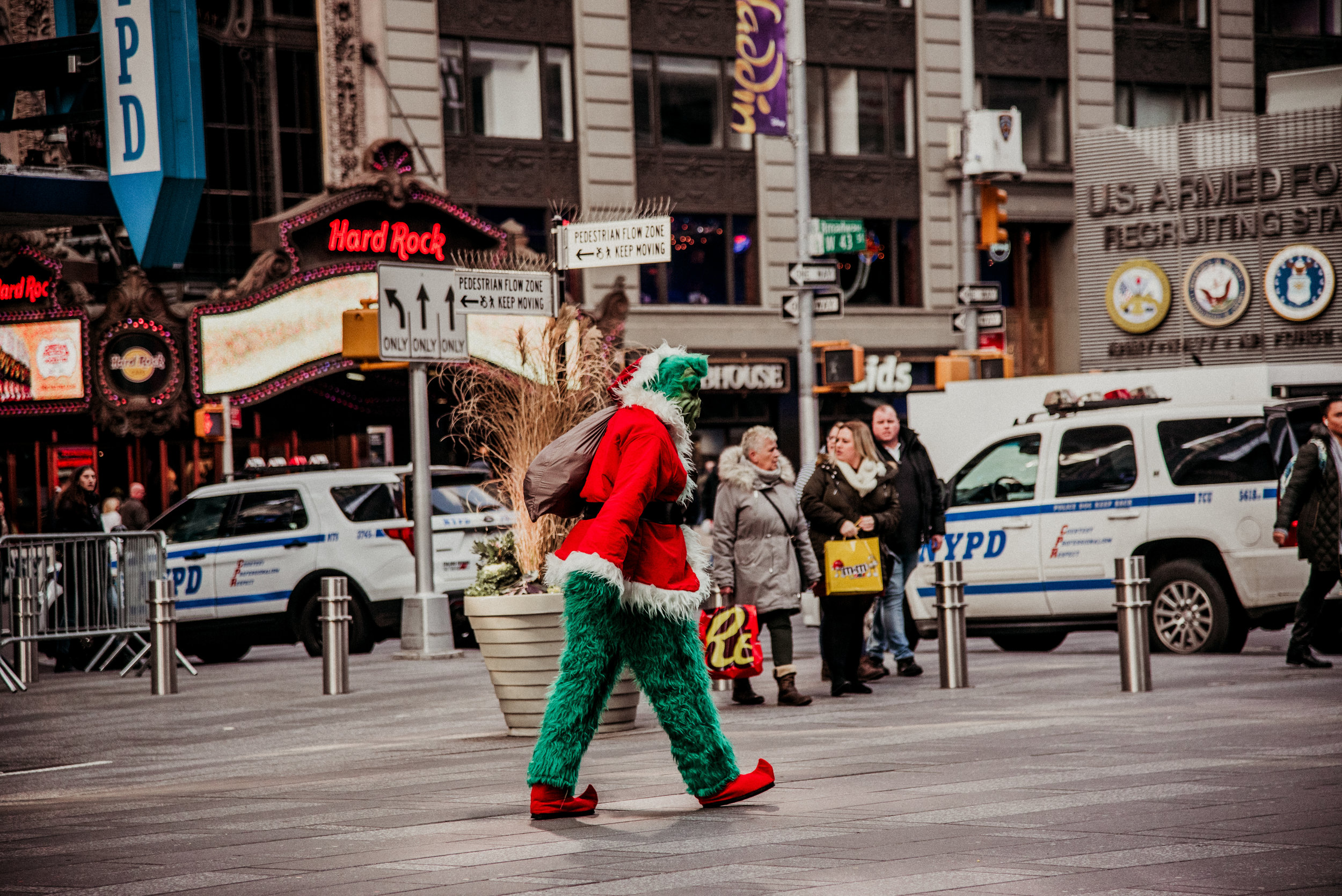 A person dressed as the Grinch in a red and green Christmas outfit walking across a busy city street at night. The background features neon signs for Hard Rock Cafe, police cars, and other pedestrians.