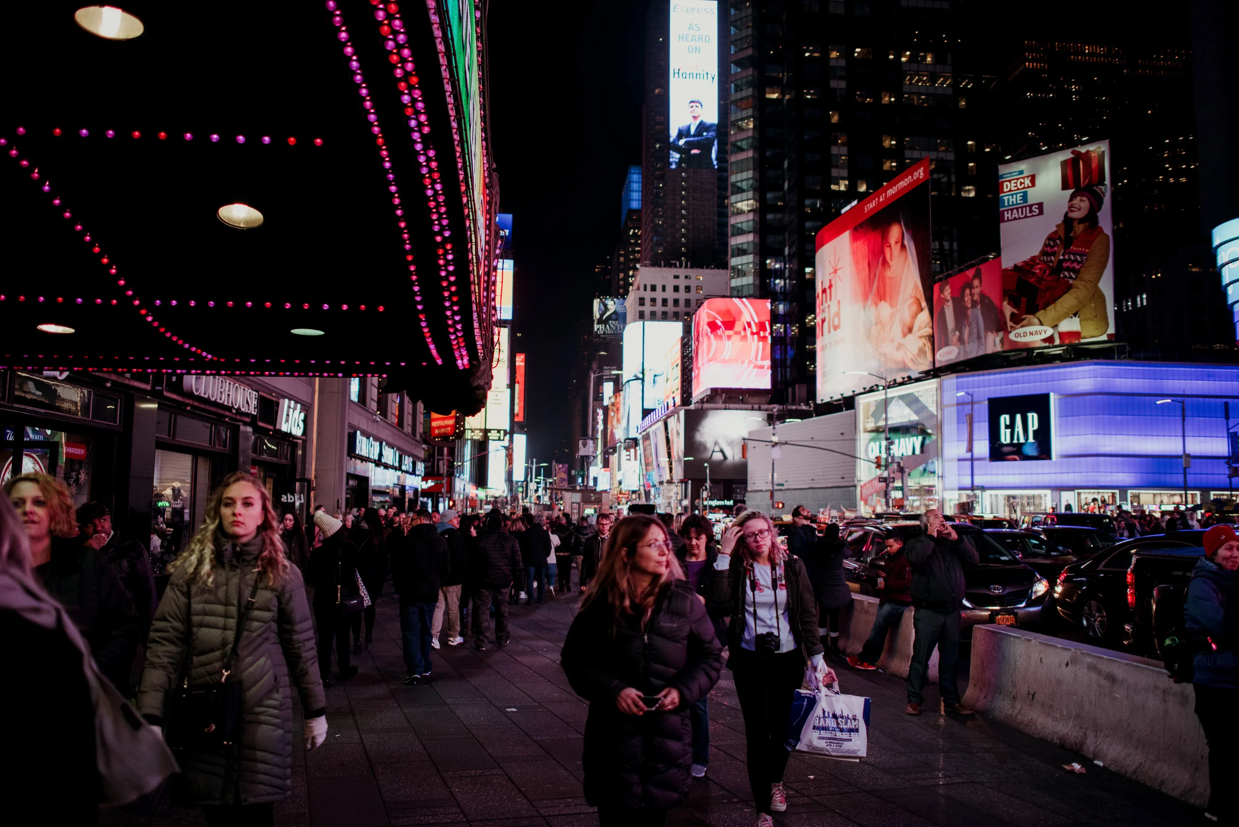 Night scene of Times Square with bright digital billboards, a crowd of pedestrians walking along the sidewalk, and illuminated storefronts and advertisements.