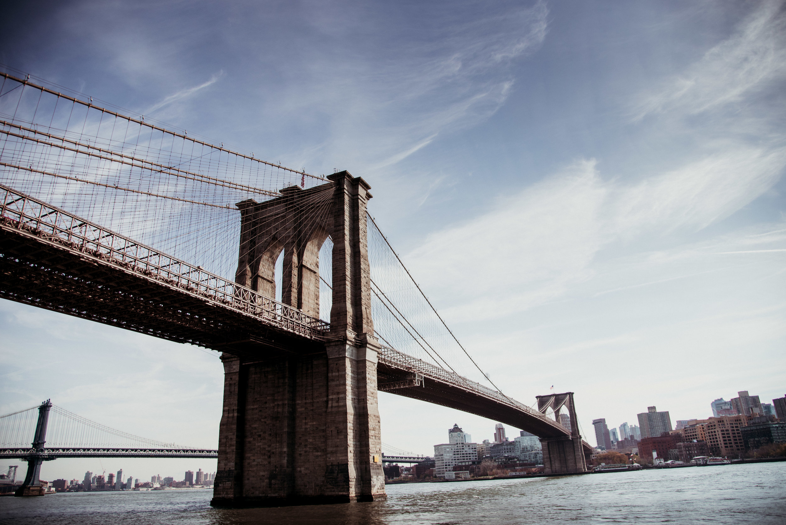 View of the Brooklyn Bridge over the East River with Manhattan skyline in the background on a cloudy day.