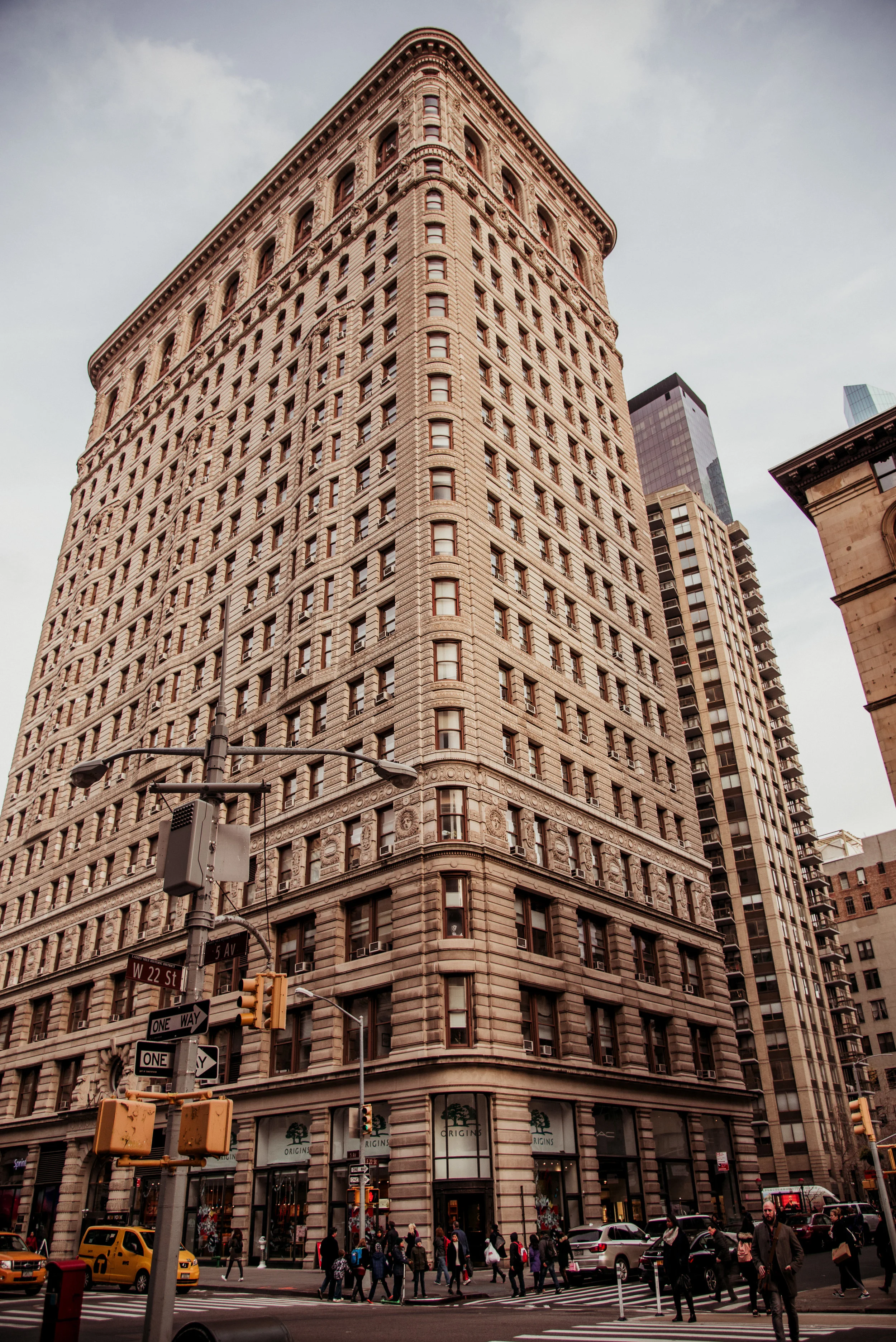 Tall historic building in New York City with multiple floors, shops at street level, and people crossing the street.