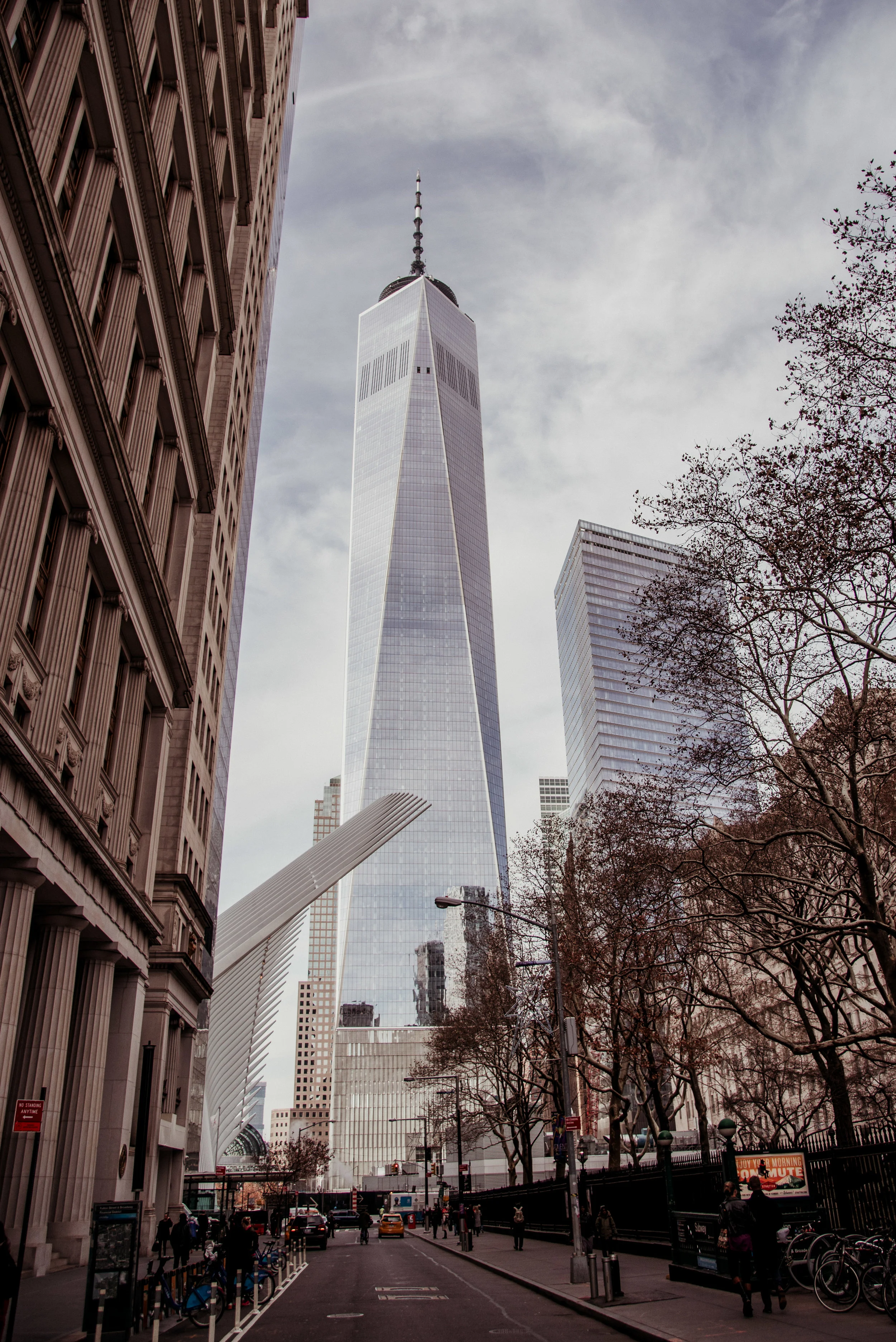 A street view of One World Trade Center in New York City surrounded by skyscrapers and leafless trees, with people walking on the sidewalk.
