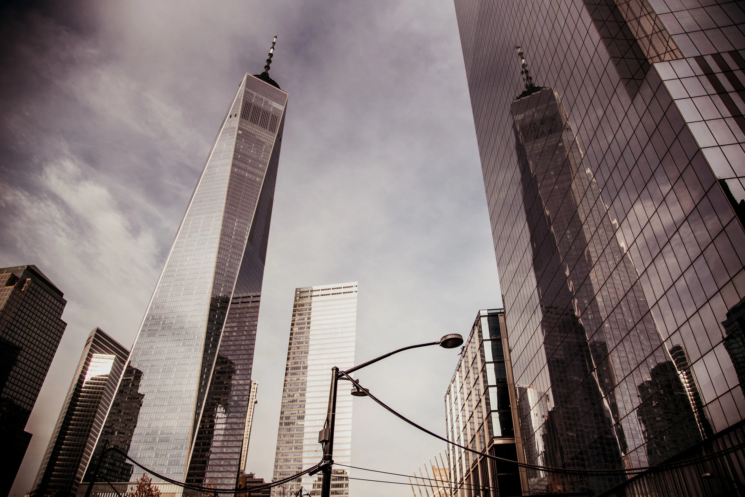 Skyscrapers in lower Manhattan, including One World Trade Center, with a partly cloudy sky and city street view in the foreground.