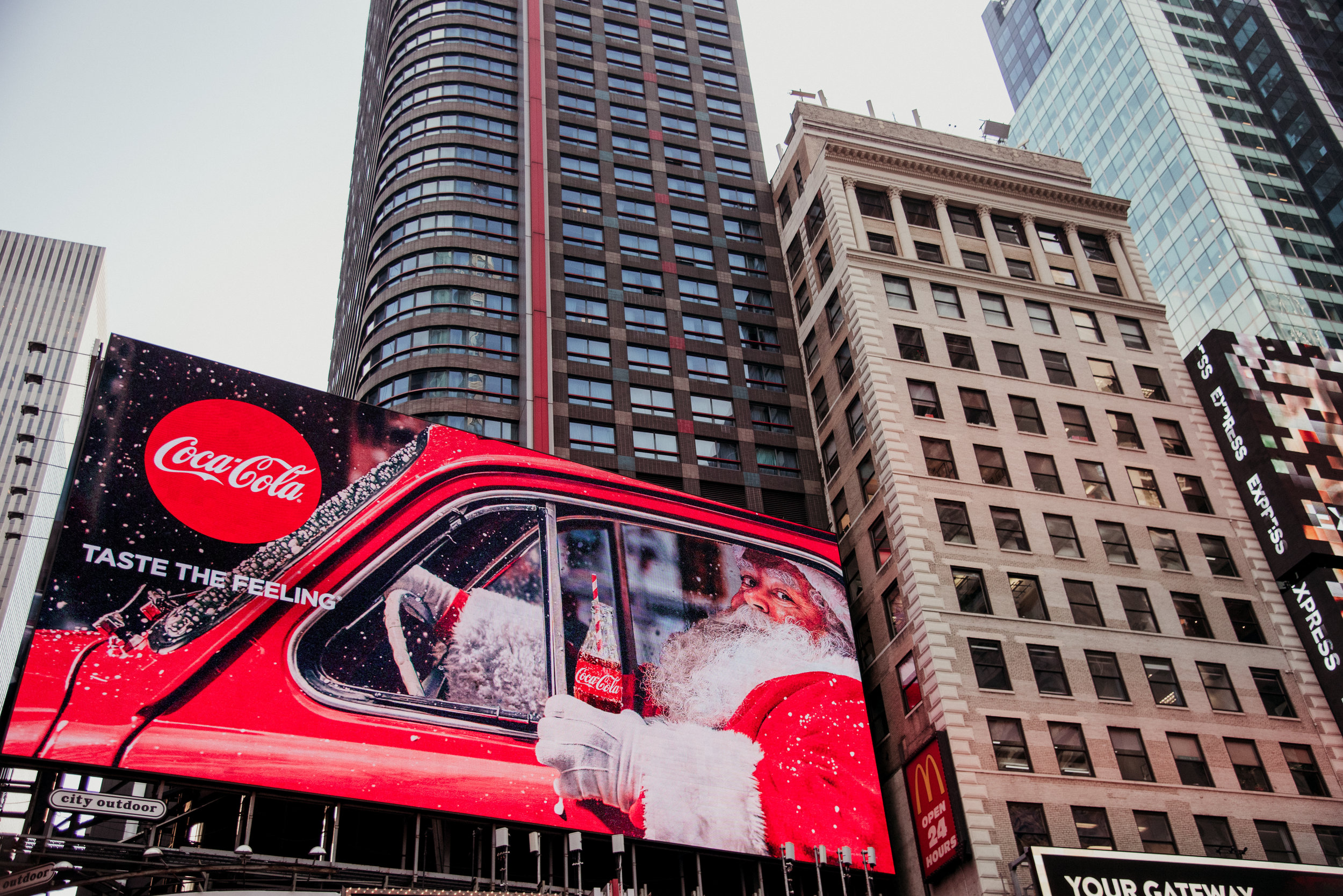 Santa Claus in Times Square holding a Coca-Cola bottle, with a large Coca-Cola billboard and tall buildings in the background.