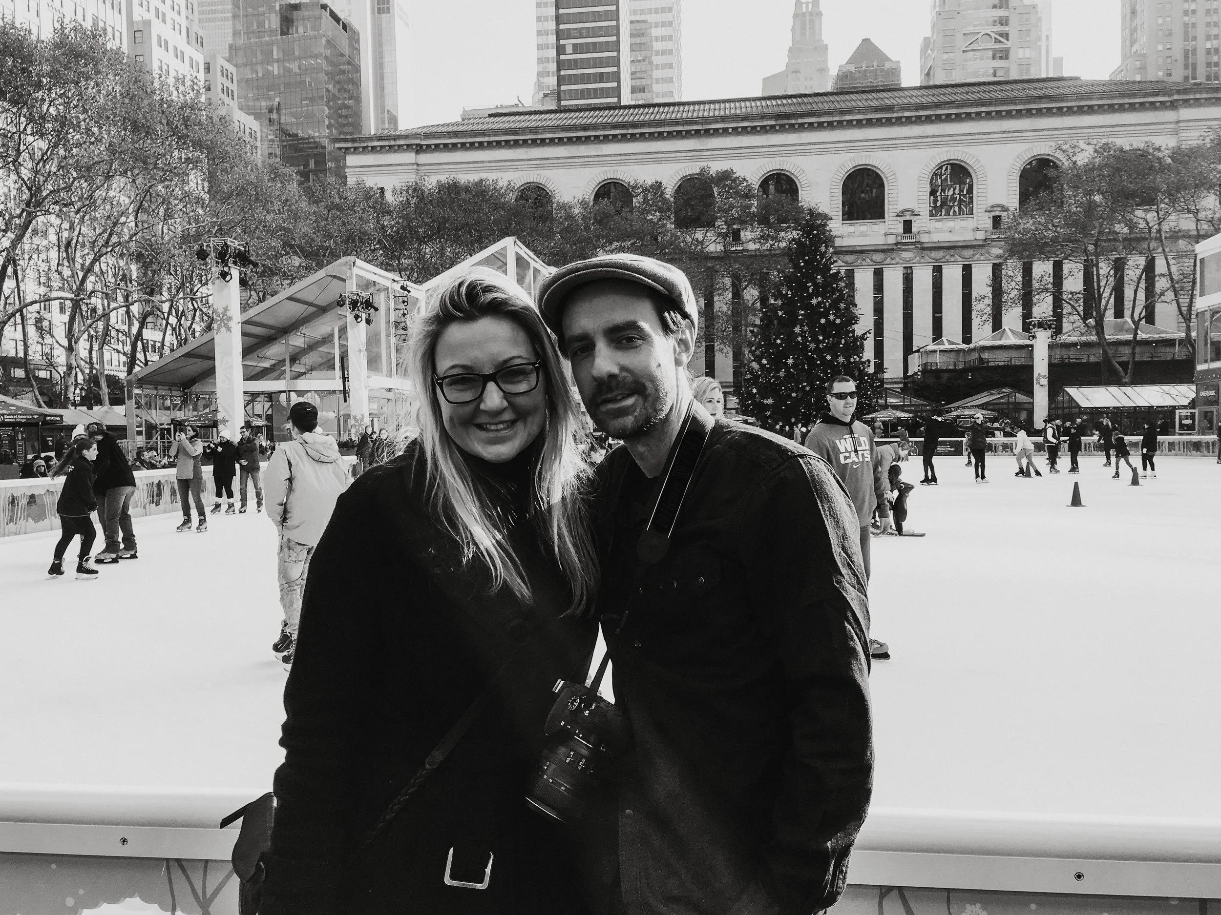 Two people smiling and posing for a photo on an outdoor ice skating rink in a city park with a Christmas tree and tall buildings in the background.