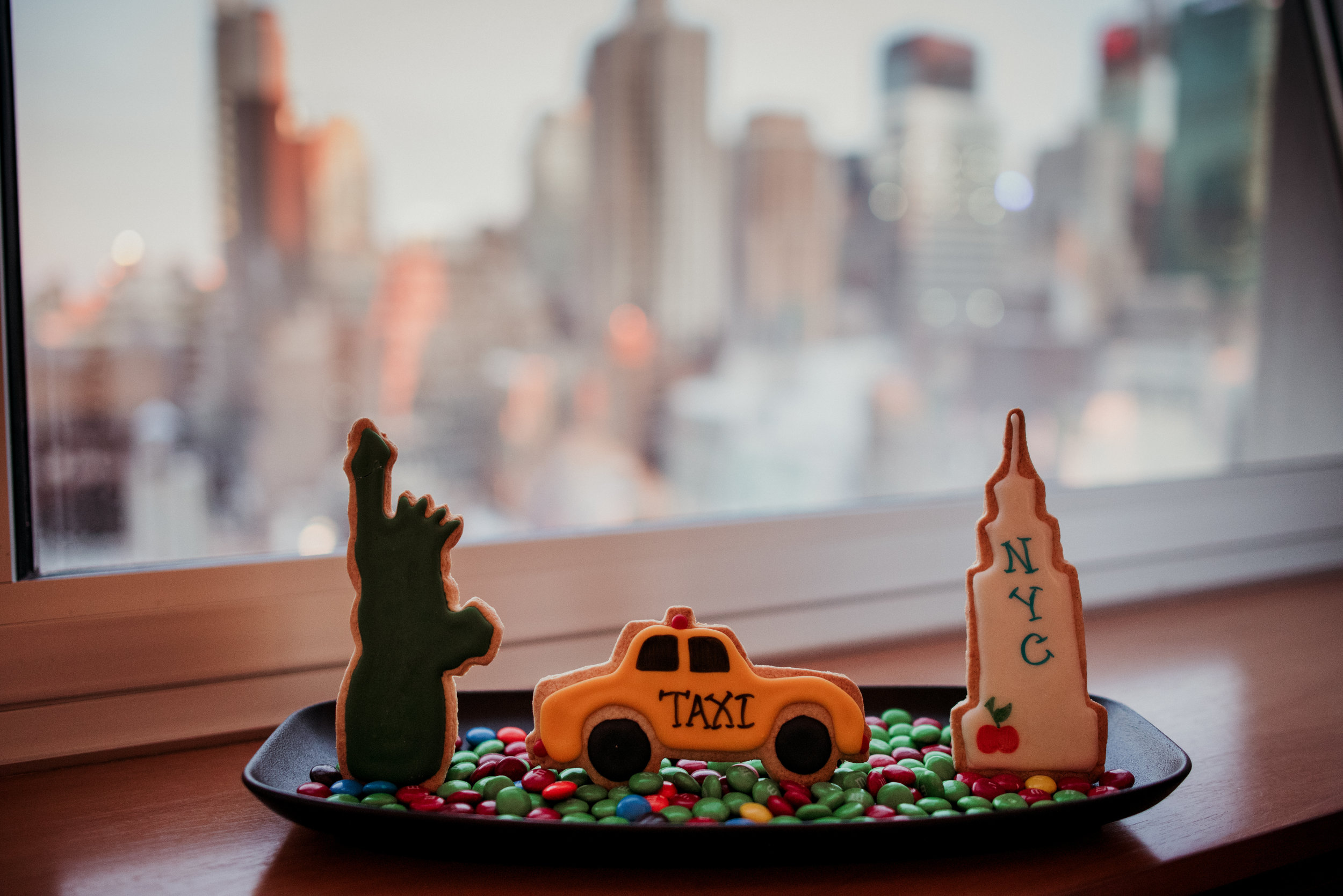 Decorative cookies with New York City theme, including a green Statue of Liberty cookie, a yellow taxi cookie, and a tall New York City landmark cookie with cherry decoration, placed on a black tray with colorful candy, against a blurred city skyline