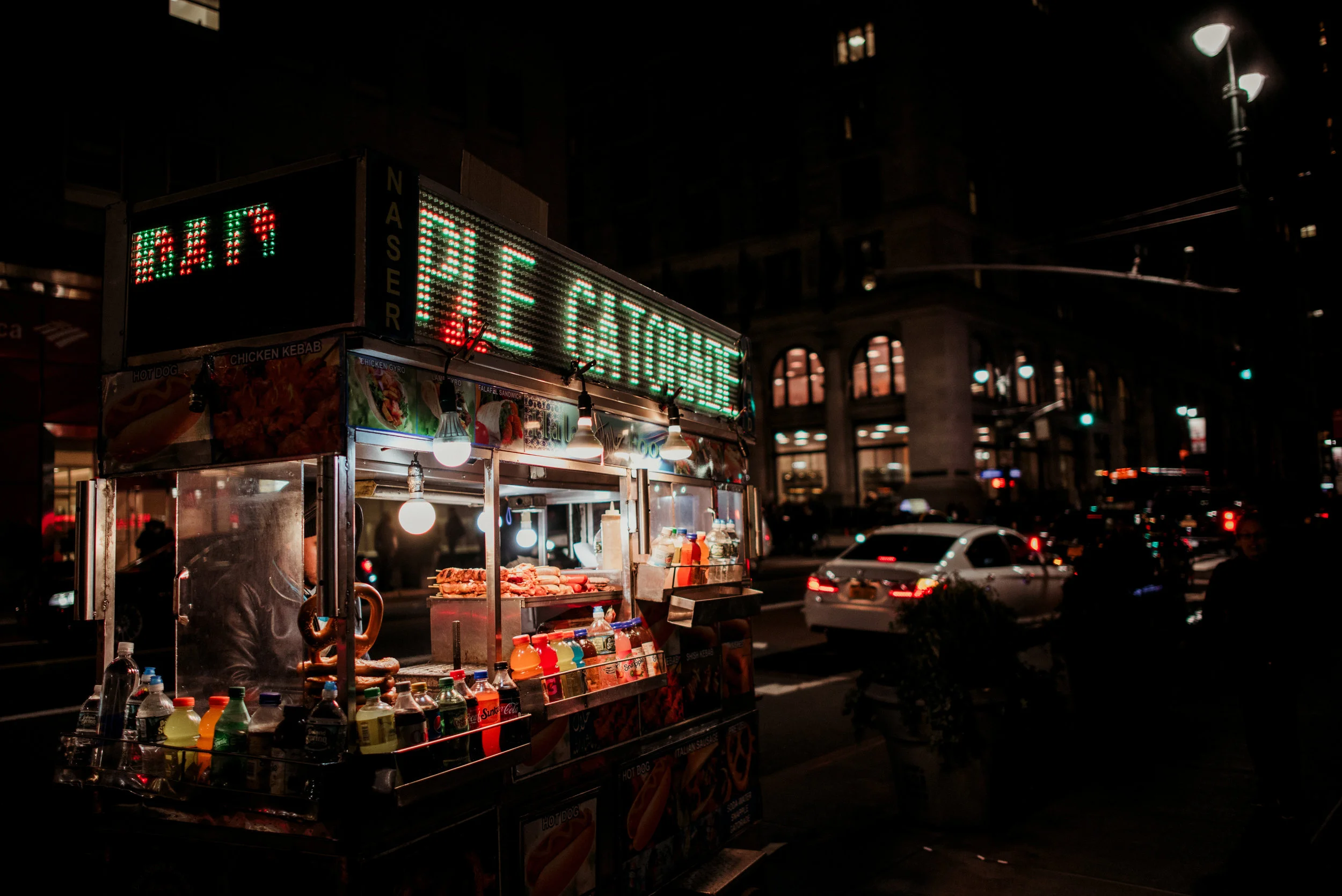 Nighttime city street scene with a lit street food cart selling hot dogs, beverages, and snacks, illuminated by hanging bulbs, with illuminated signs and cars in the background.